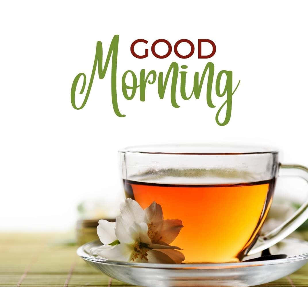 寧靜晨光中的溫暖茶杯 壁紙 — 一個透明玻璃茶杯裝滿琥珀色茶，放在碟子上，旁邊有一朵白花，背景是綠色竹蓆，並有「GOOD Morning」文字。