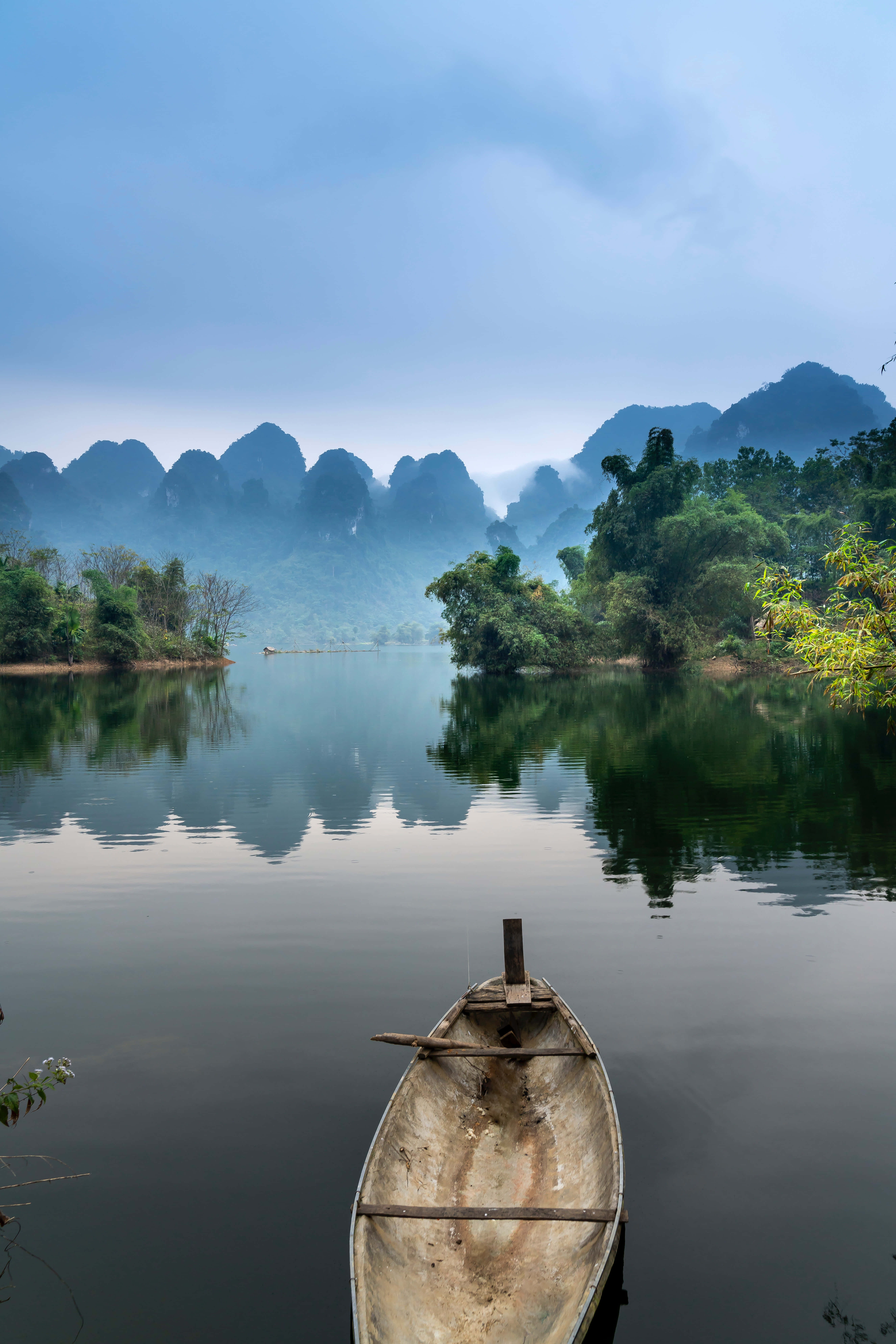 寧靜河上的小船 壁紙 — 一艘風化的木船漂浮在玻璃般的河面上，映照著霧氣繚繞的喀斯特山脈，天空柔和湛藍。