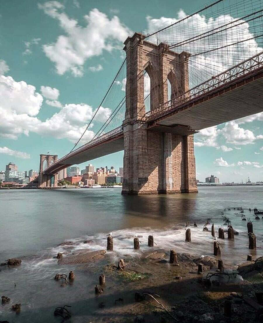 寧靜河面的日落美景，遠處可見橋樑 壁紙 — 布魯克林大橋橫跨東河，天空明亮，背景是城市天際線，前景是風化的樁柱。