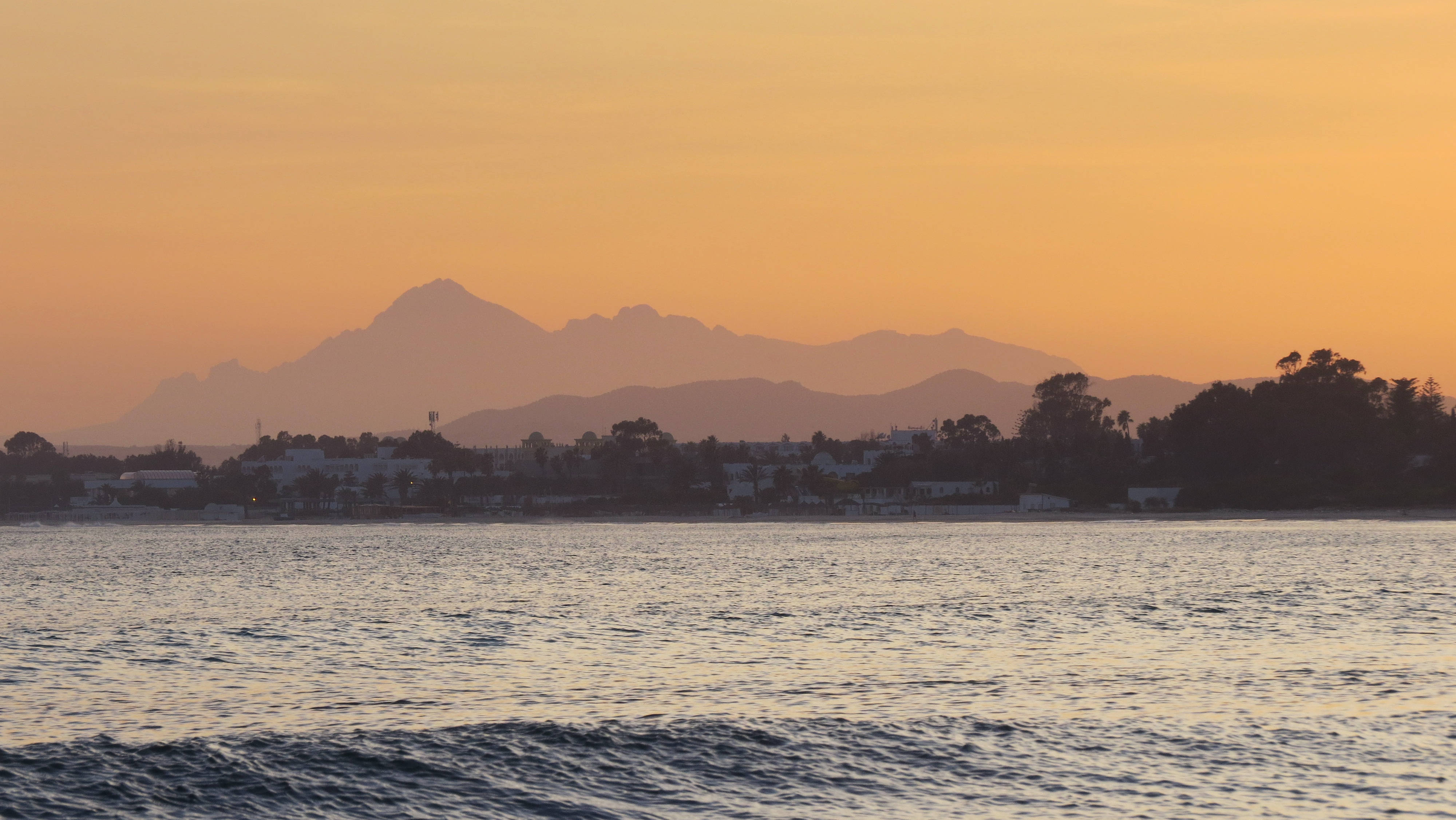 寧靜海景與山脈 壁紙 — 金色夕陽在平靜的海面上投射出溫暖的光芒，遠處有山巒剪影和沿海城鎮的輪廓。