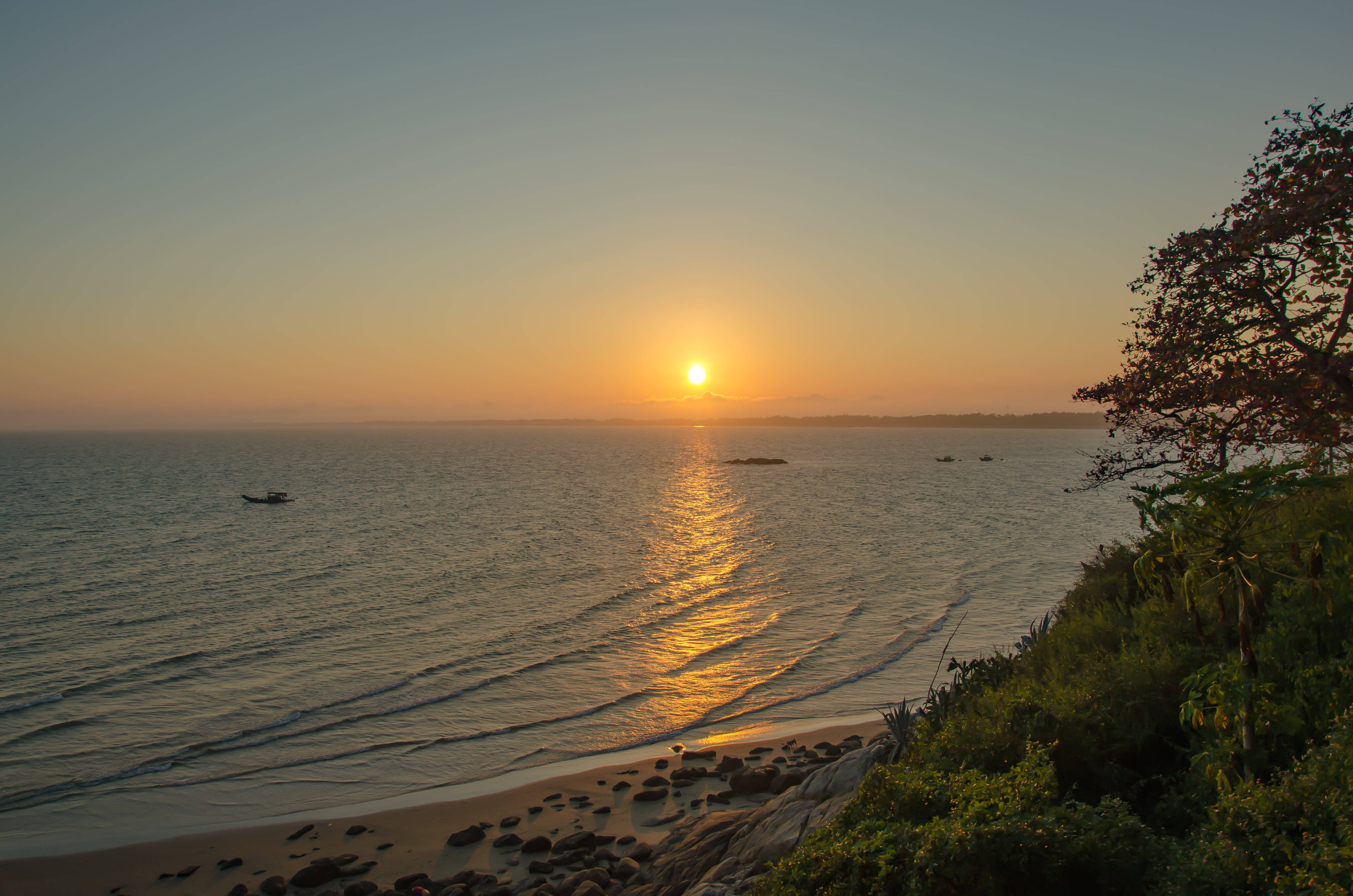 寧靜海灘日落 壁紙 — 金色夕陽照耀在平靜的海面上，小船在綠意盎然的岩石海岸附近漂流。