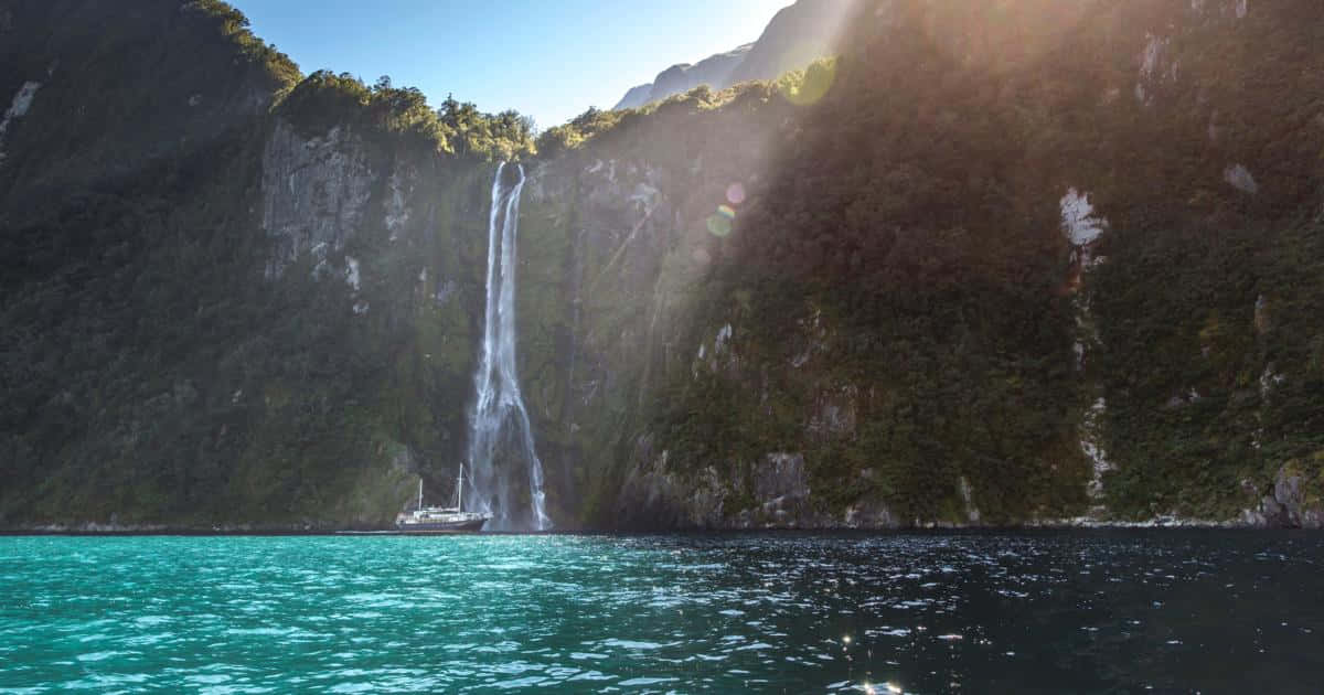 寧靜海灘與清澈海水 壁紙 — 一道雄偉的瀑布從鬱鬱蔥蔥的綠色懸崖傾瀉而下，流入碧綠的海水中，下方有一艘小船，陽光穿透薄霧。