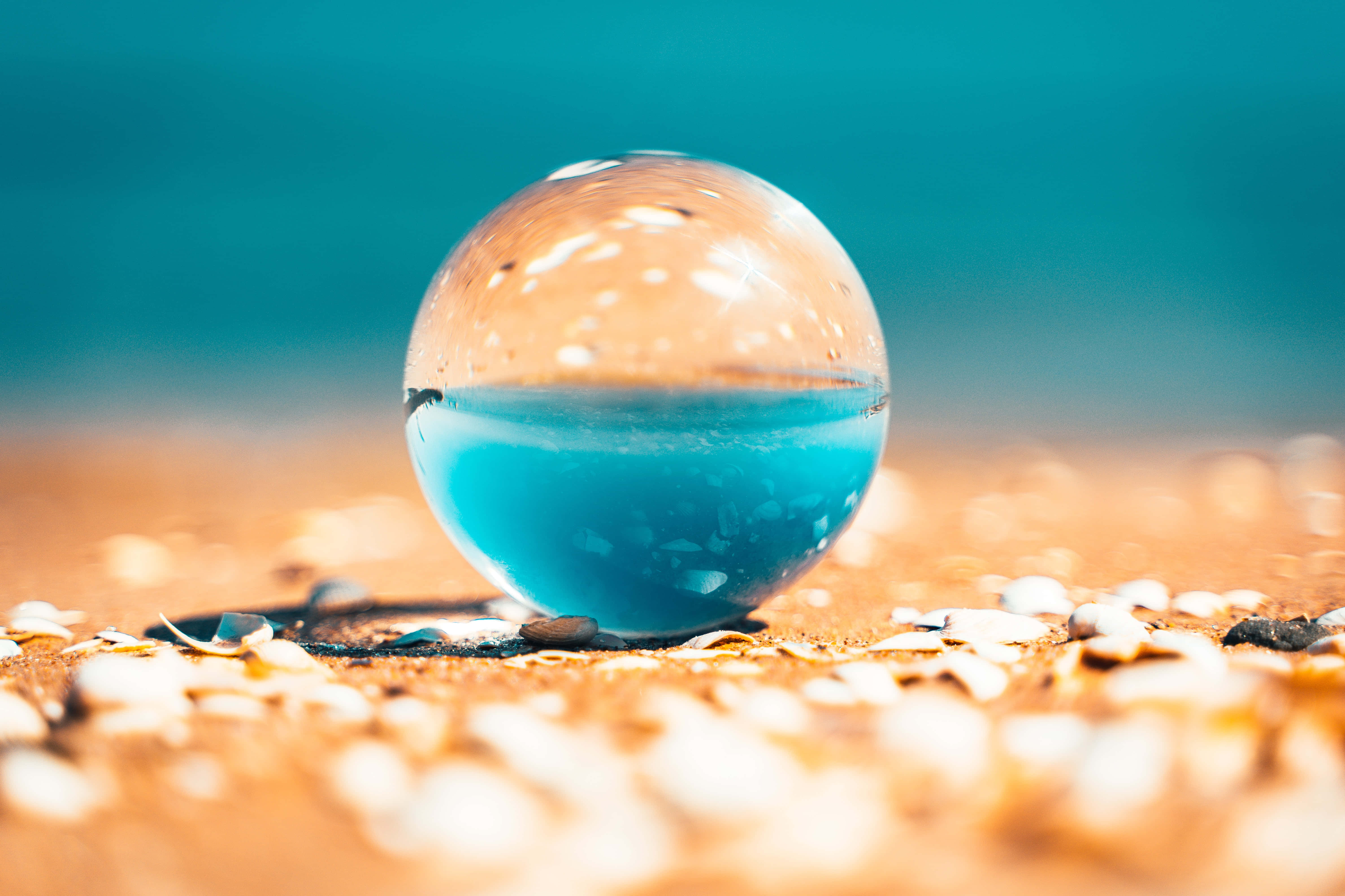 寧靜海面的壯麗日落 壁紙 — 一個透明玻璃球充滿鮮豔藍色液體，放在沙灘上，背景有貝殼和柔焦的碧綠海洋。