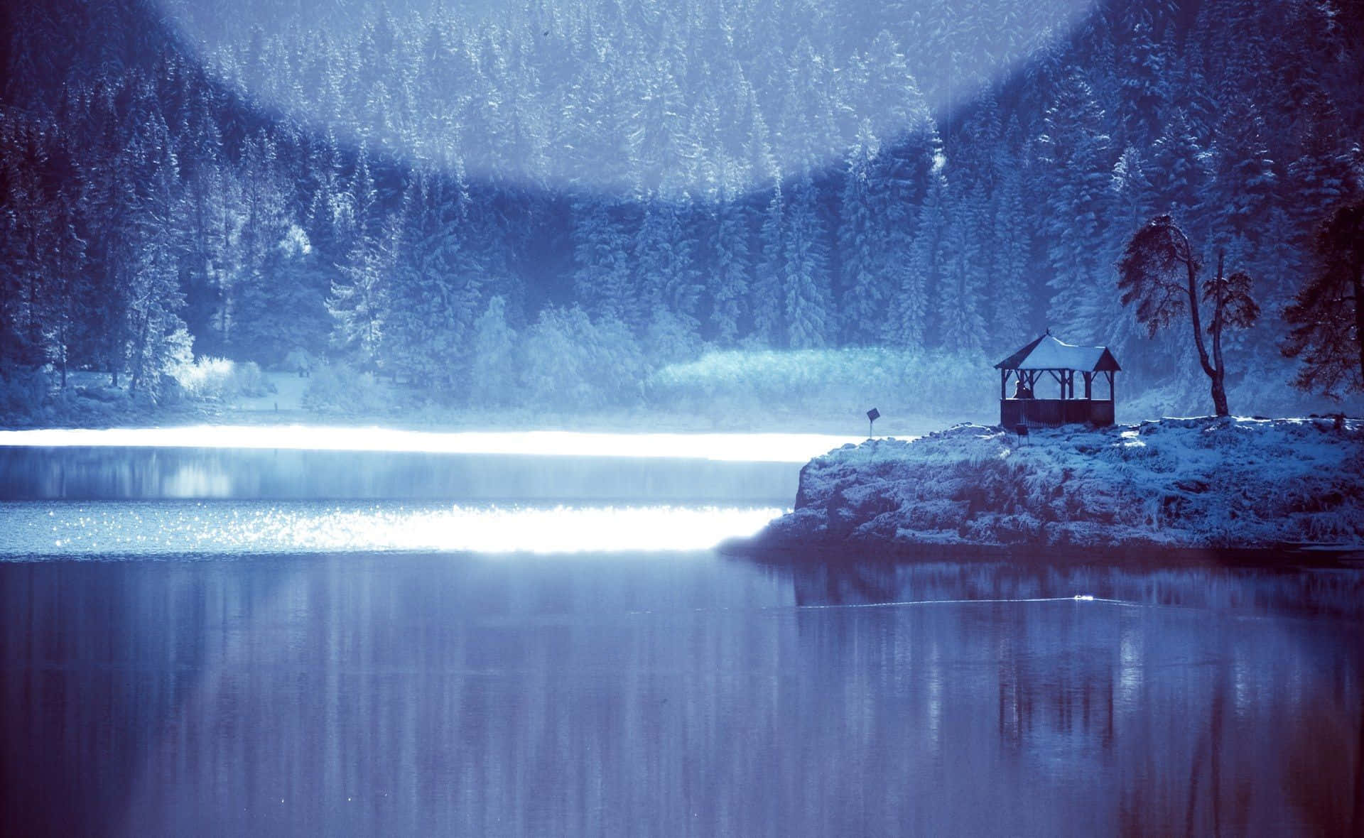 Frozen Lake 桌布 壁紙 — 寧靜的冬日湖泊倒映著白雪覆蓋的森林和一座孤獨的木製涼亭，在閃耀的藍天下。