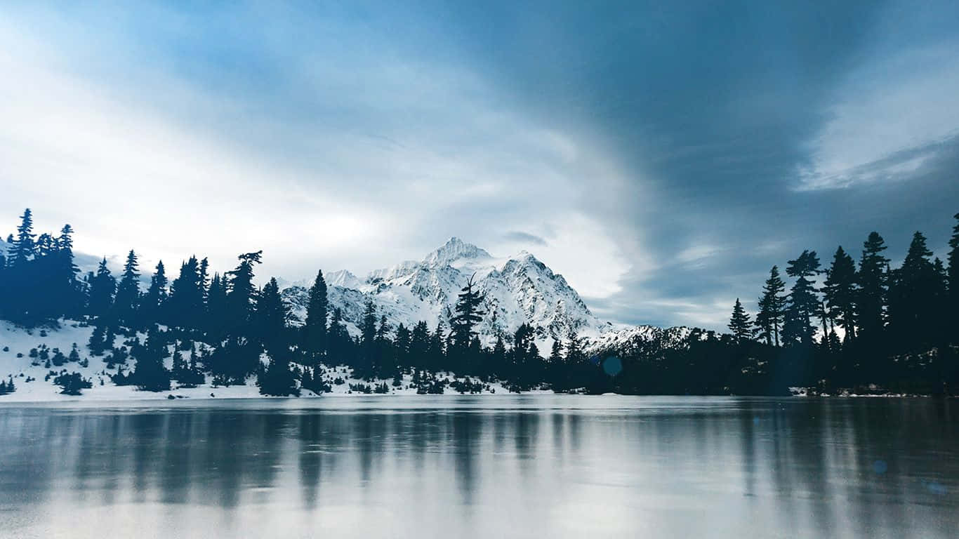 Frozen Lake 桌布 壁紙 — 寧靜的冬日湖面映照著積雪的山峰和常綠樹，天空憂鬱的藍色。