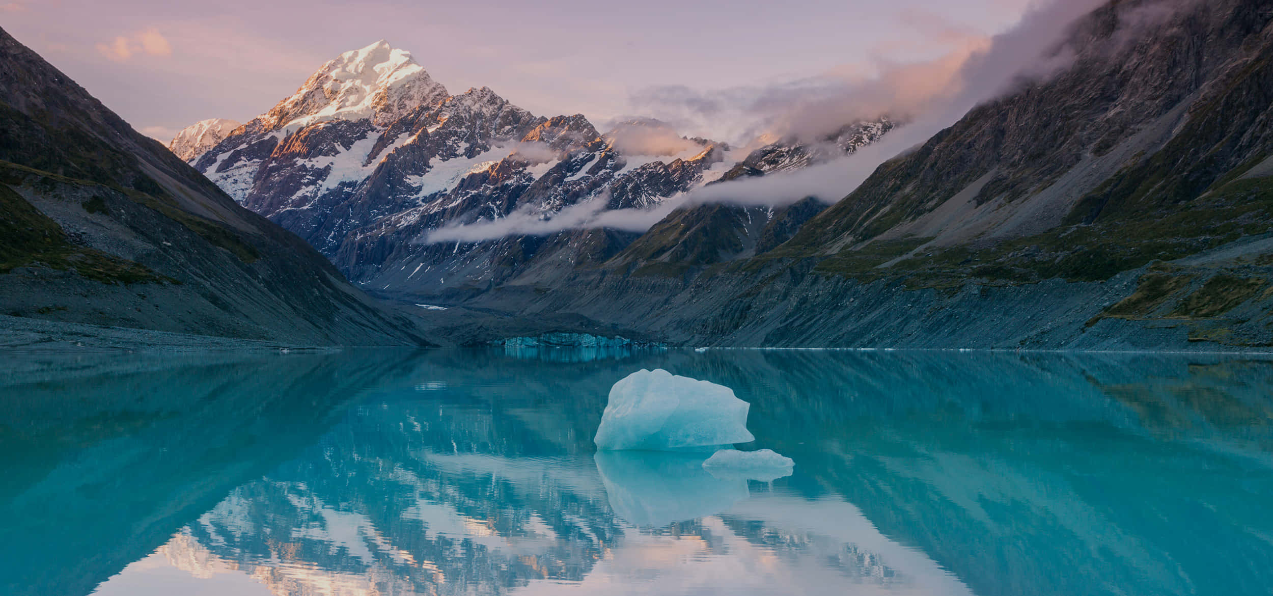 New Zealand 桌布 壁紙 — 寧靜的冰川湖在黎明時分映照著白雪覆蓋的山脈和柔和的粉紅雲彩，並有一座漂浮的冰山。