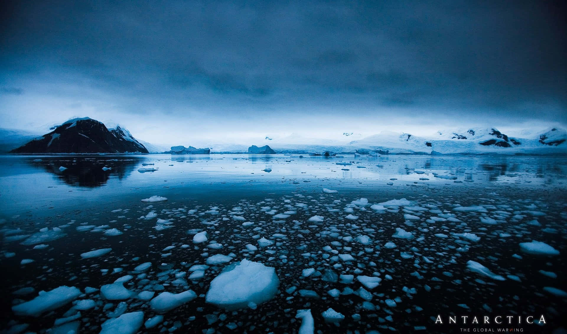 Antarctica 桌布 壁紙 — 寧靜的南極場景，冰凍的水面上漂浮著冰塊，積雪覆蓋的山脈在陰鬱的藍天下。