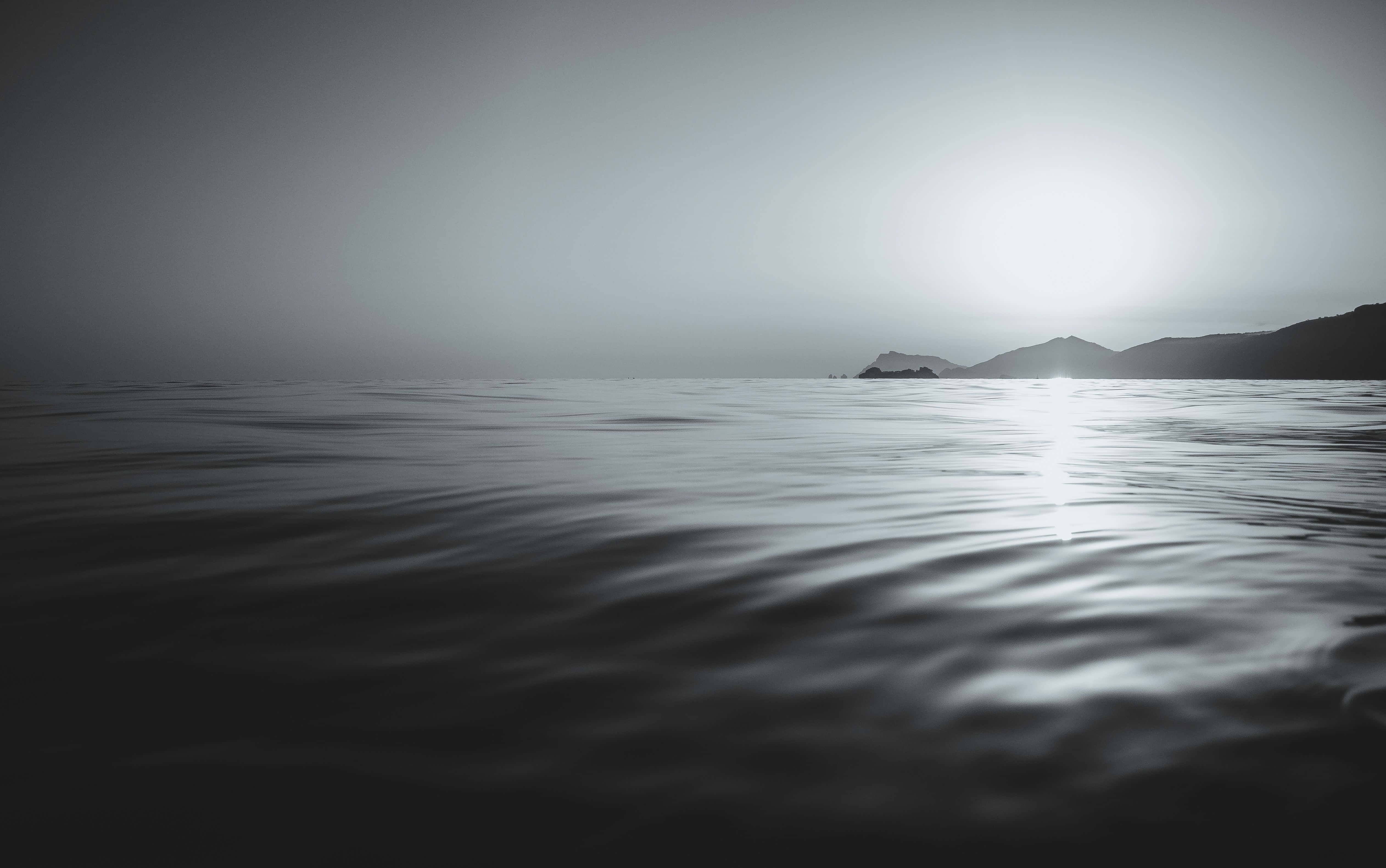 Black And White Ocean 桌布 壁紙 — 寧靜的單色海景，柔和漣漪映照出明亮太陽，遠方山丘輪廓分明。
