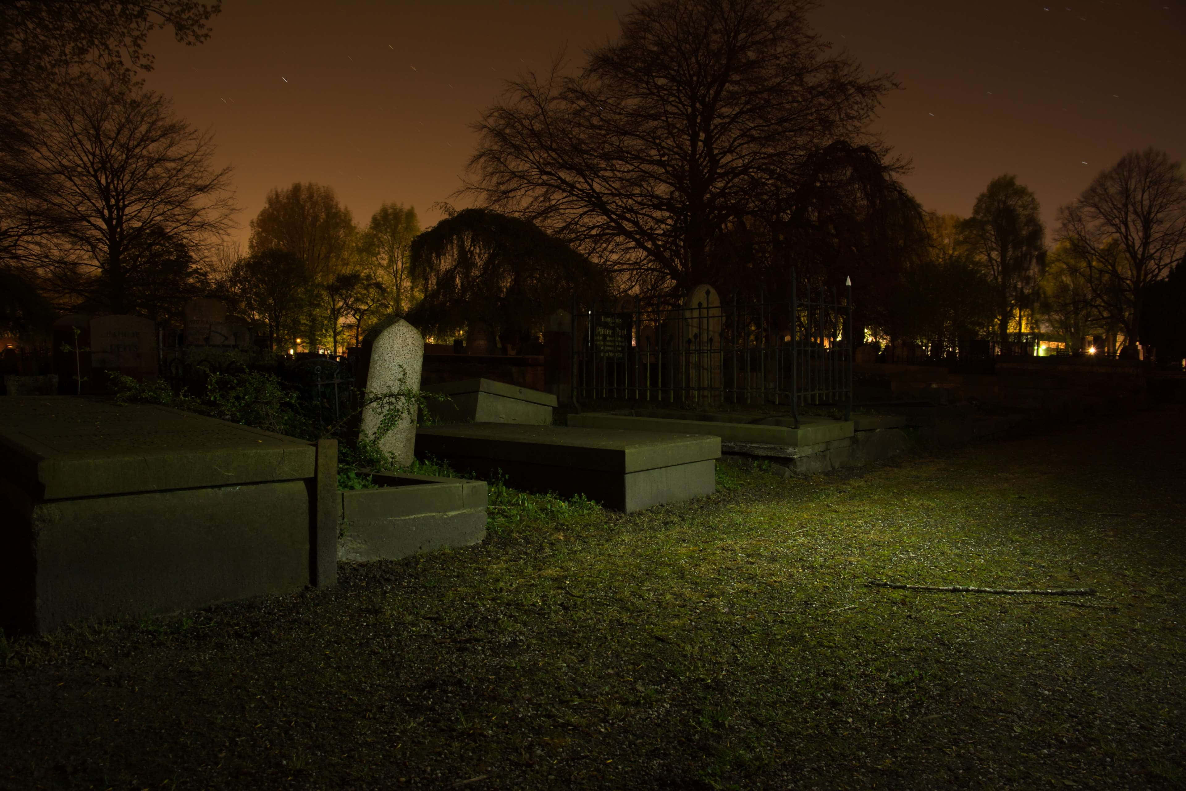 Gravestone 桌布 壁紙 — 寧靜的夜晚墓地，墓碑在滿是星光的橙色天空下，光禿的樹木在遠方燈光映襯下形成剪影。