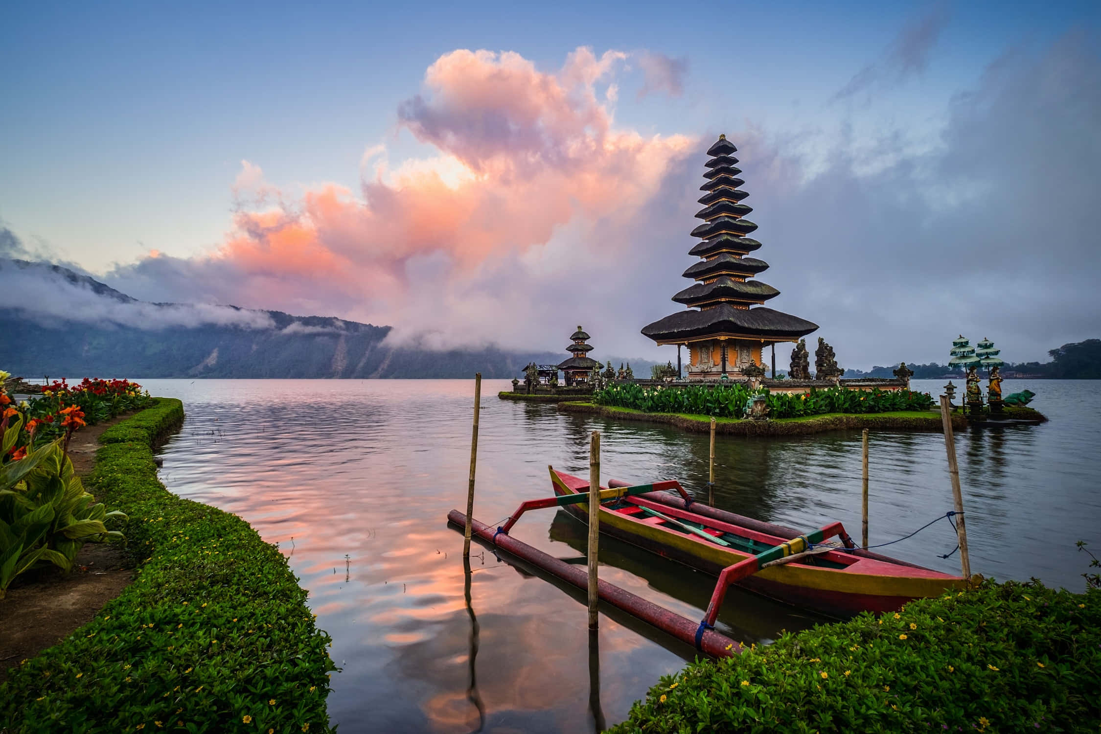 Indonesia 桌布 壁紙 — 寧靜的日出，映照在平靜的湖面上，湖中小島上有一座傳統的峇里島寺廟，附近停泊著色彩繽紛的船隻。