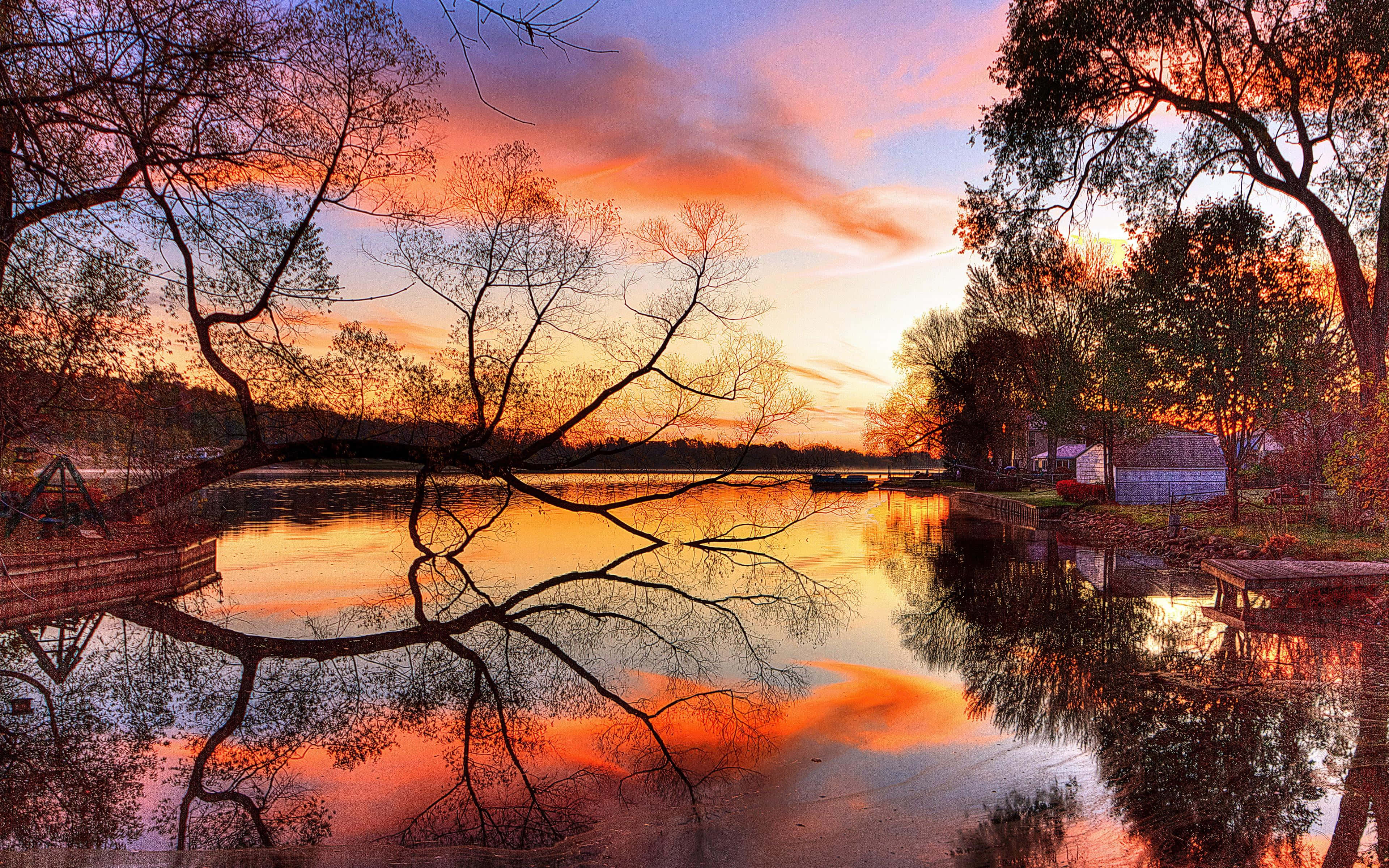 Sunset Reflection 桌布 壁紙 — 寧靜的日落映照在平靜的湖面上，天空呈現鮮豔的橙色和粉紅色，倒映在水中，周圍是光禿禿的樹木和一個碼頭。