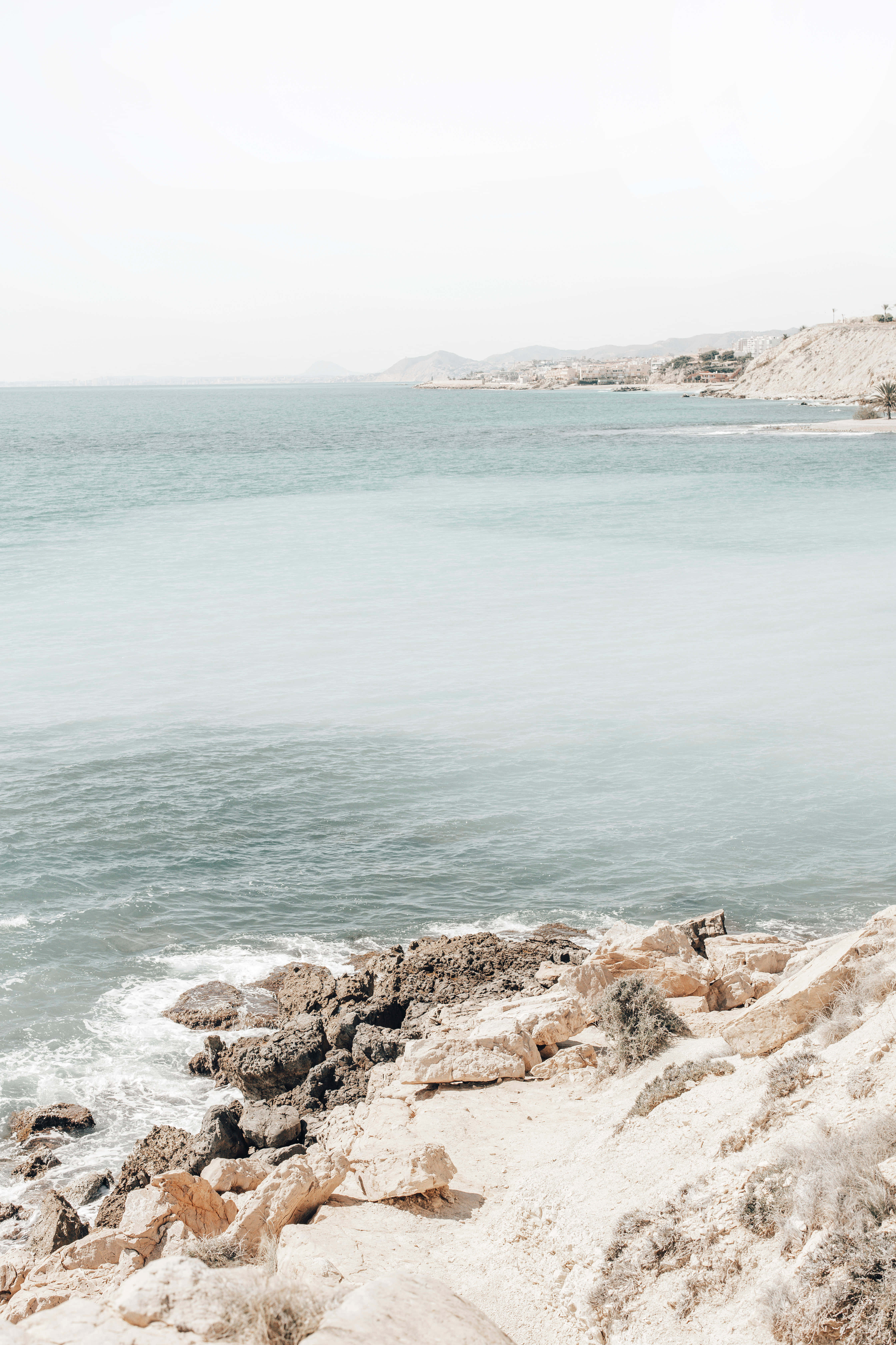Mediterranean Beach 桌布 壁紙 — 寧靜的海岸景色，平靜的碧綠海水在朦朧的天空下拍打著陽光普照的岩石海岸。