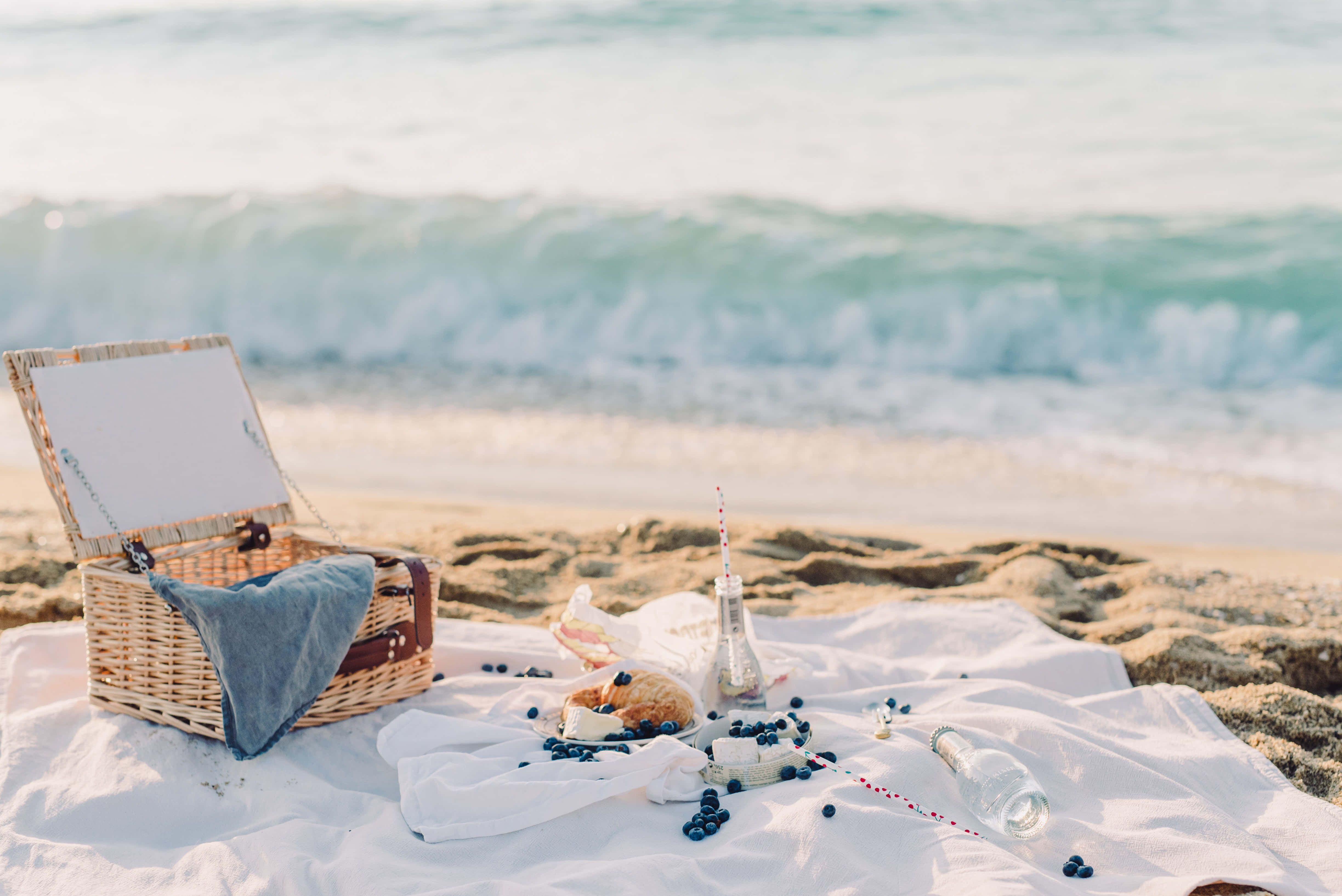 Beach Picnic 桌布 壁紙 — 寧靜的海灘野餐，配有柳條籃、藍色毯子和藍莓，背景是溫柔的海浪。