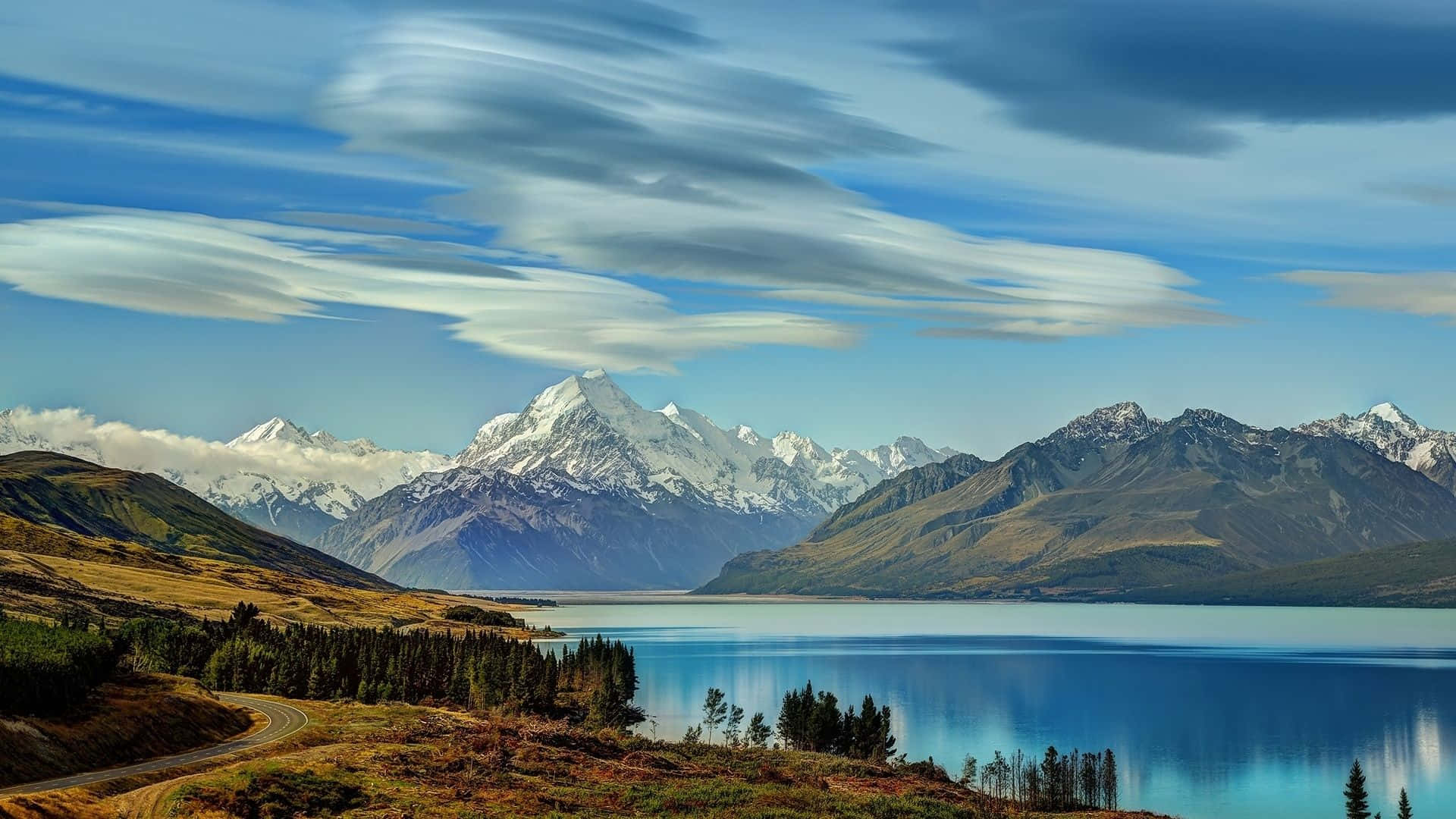 New Zealand 桌布 壁紙 — 寧靜的湖面映照著積雪的山峰，天空中有條紋狀的雲彩，沿岸有一條蜿蜒的道路。