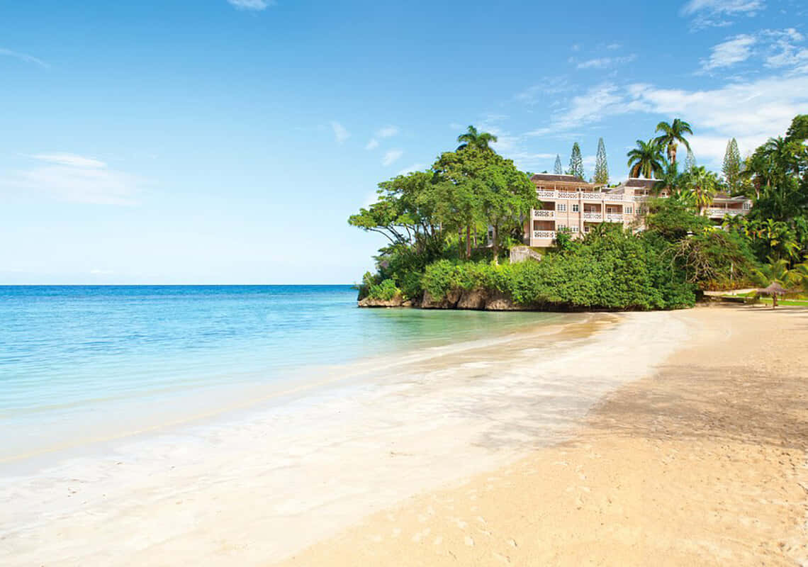 Jamaican Beach 桌布 壁紙 — 寧靜的熱帶海灘，碧綠的海水輕輕拍打金色沙灘，旁邊是綠意盎然的島嶼，島上有度假村建築。