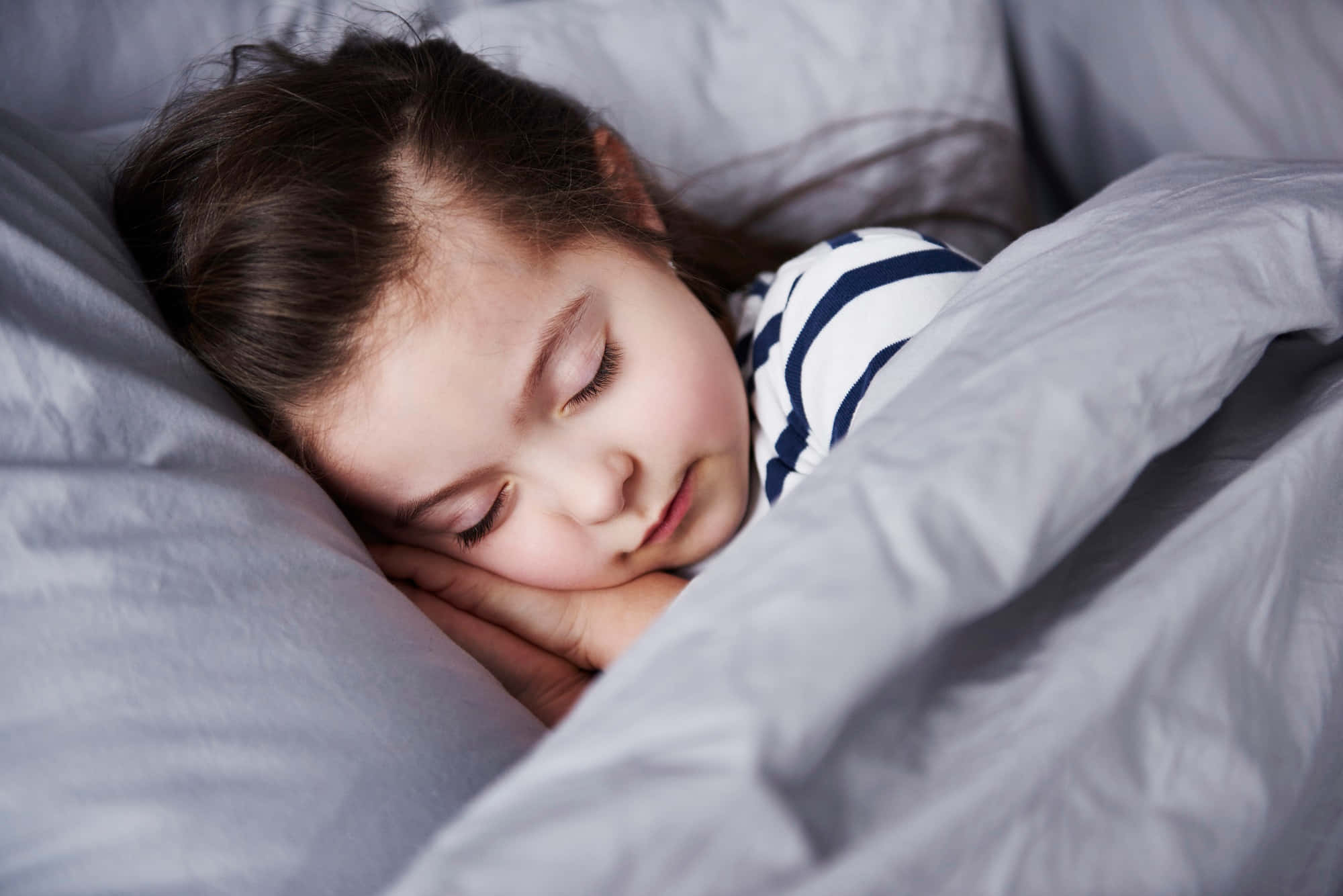 寧靜的睡眠 壁紙 — 一個年輕女孩將頭枕在手上，依偎在柔軟的灰色寢具中，安詳地睡著。