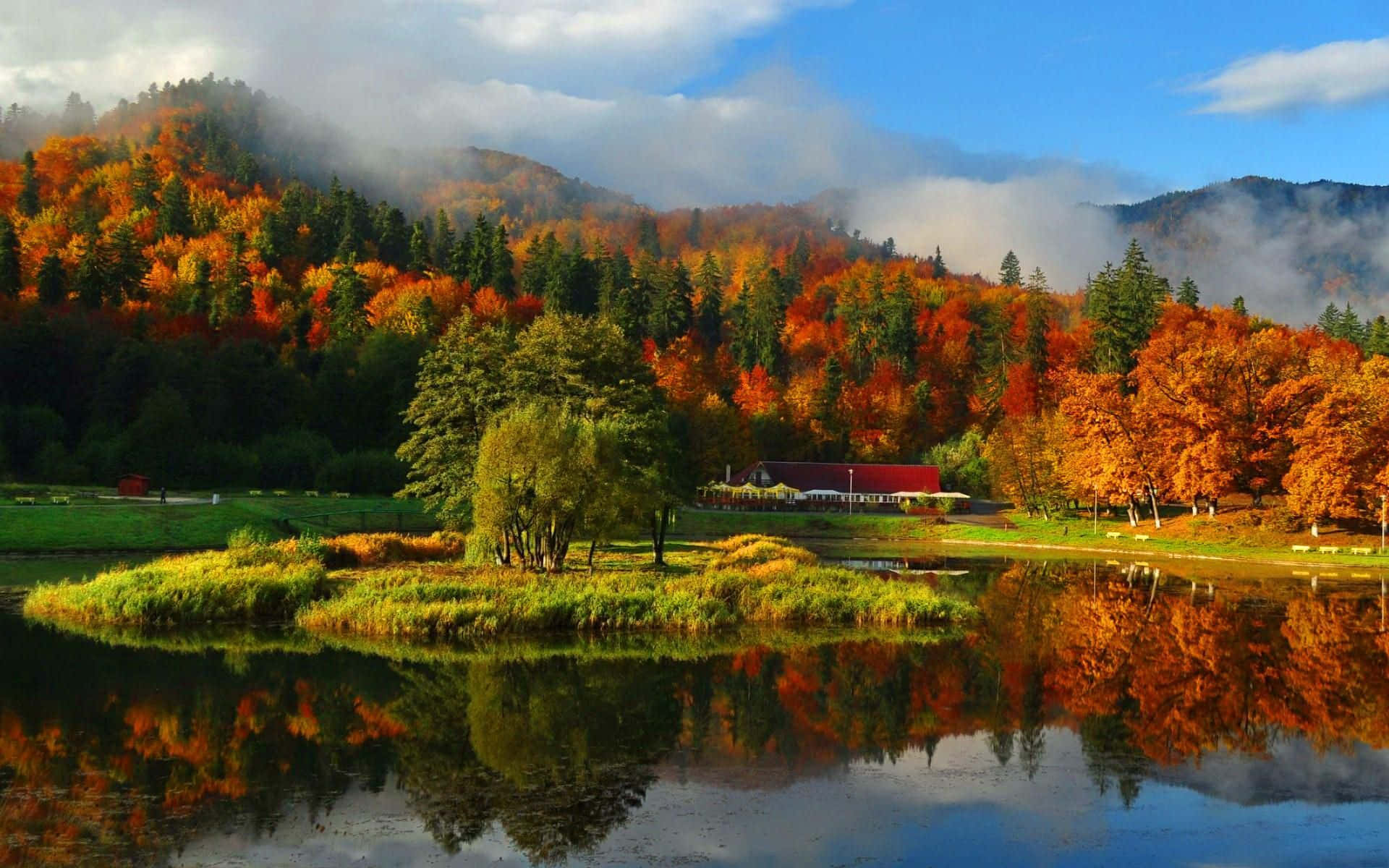 Fall Mountains 桌布 壁紙 — 寧靜的秋日湖泊映照出環繞著紅頂小屋的熾熱橙色和綠色樹木，小屋坐落在霧氣繚繞的山巒中。