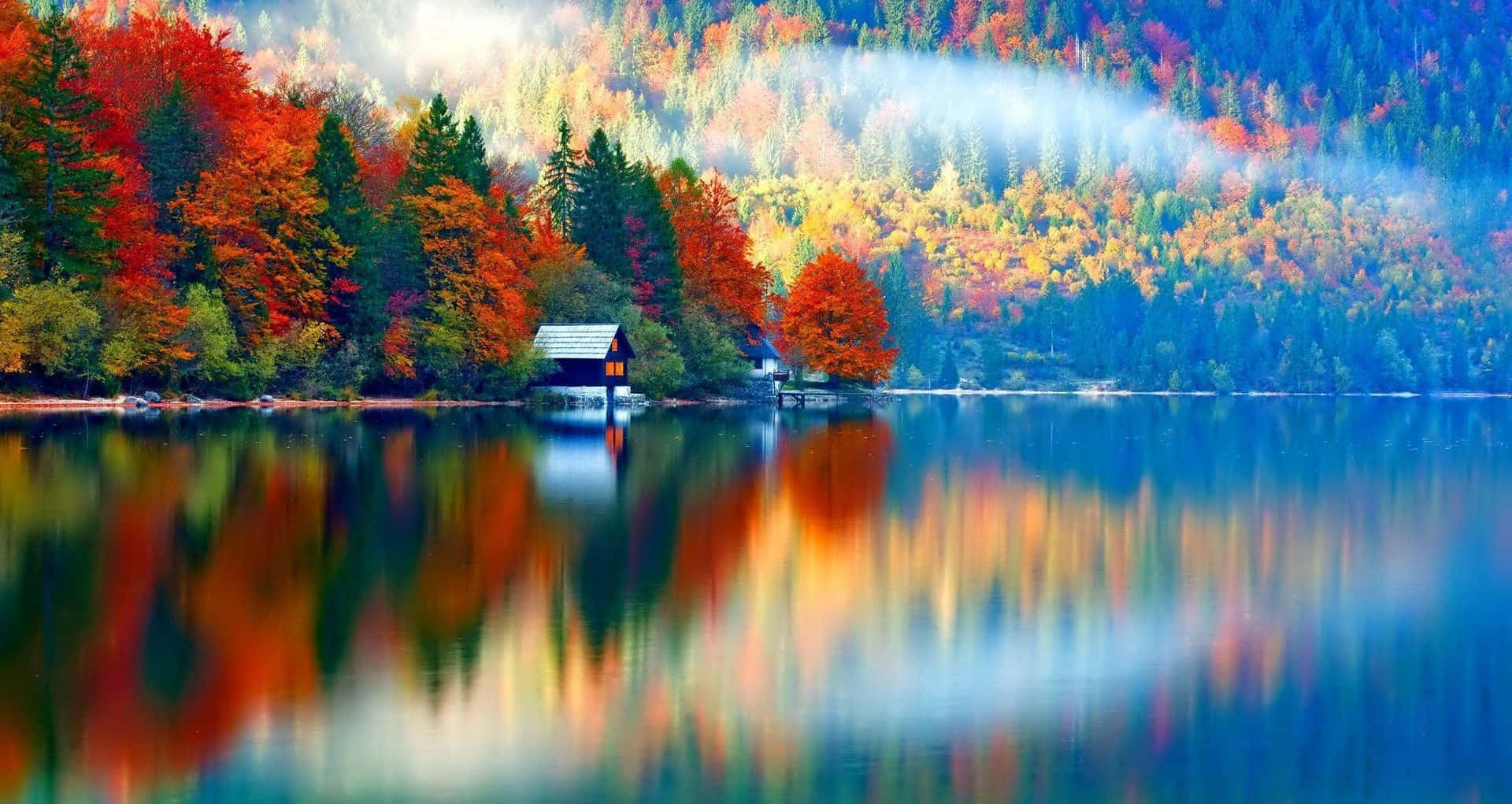 Autumn Colors 桌布 壁紙 — 寧靜的秋日湖面映照著火紅、橙黃和金黃的樹木，其間有一座小木屋坐落在霧氣繚繞的山丘下。