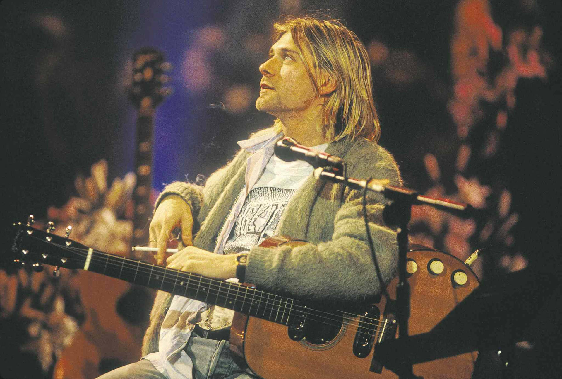 Kurt Cobain 在演唱會期間在舞台上彈奏木結他，身後有觀眾。