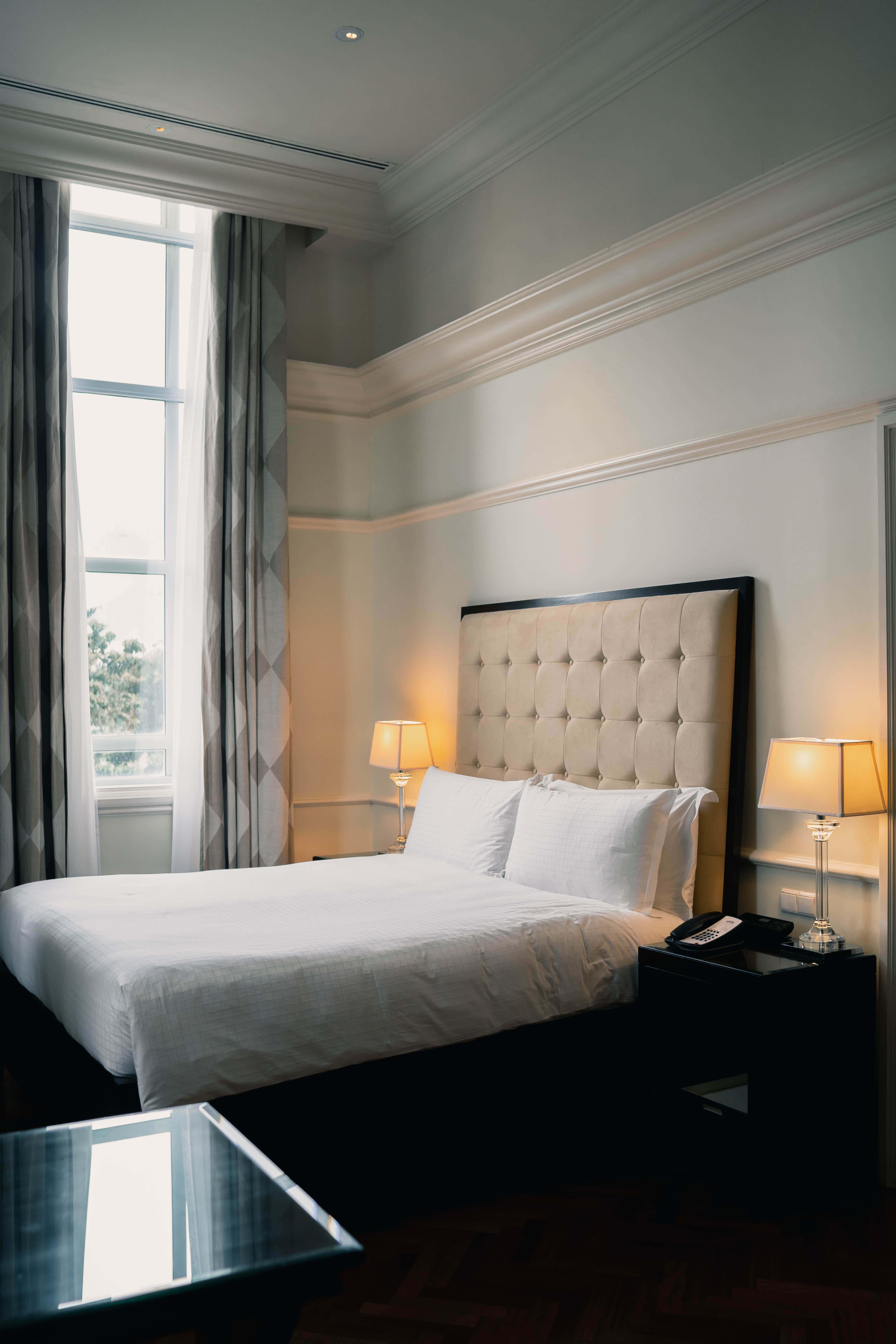 Hotel Room 桌布 壁紙 — 寧靜的酒店睡房，配有簇絨奶油色床頭板、白色床單和散發柔和光線的溫暖床頭燈。