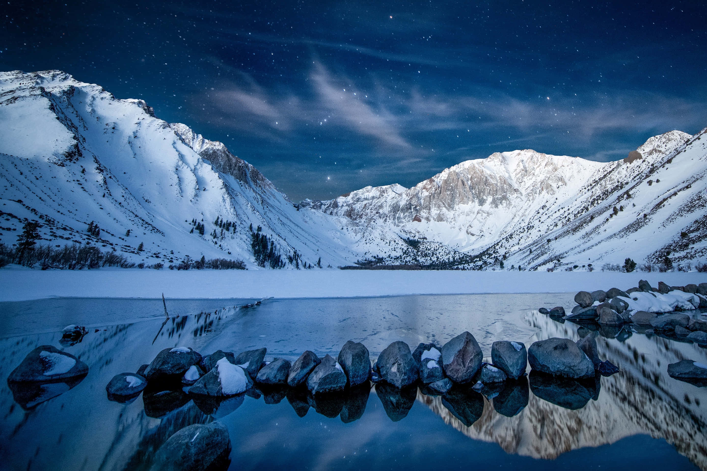 寧靜的雪山風景 壁紙 — 白雪覆蓋的山脈在繁星點點的夜空下完美地倒映在冰凍的湖面上，周圍環繞著深色的岩石。