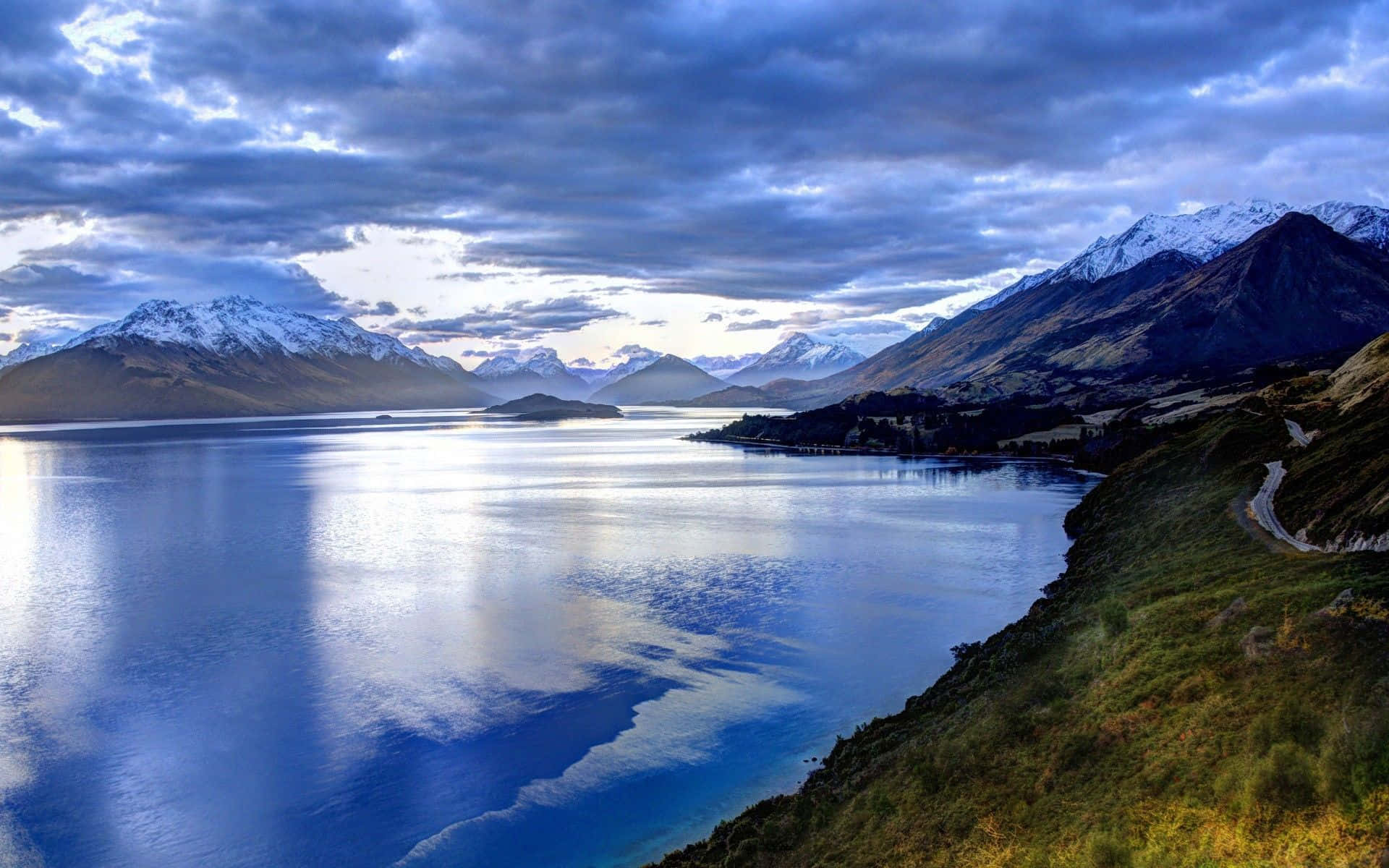 New Zealand 桌布 壁紙 — 寧靜的高山湖泊映照著白雪覆蓋的山峰，天空戲劇性地多雲，湖岸蜿蜒的道路延伸其間。