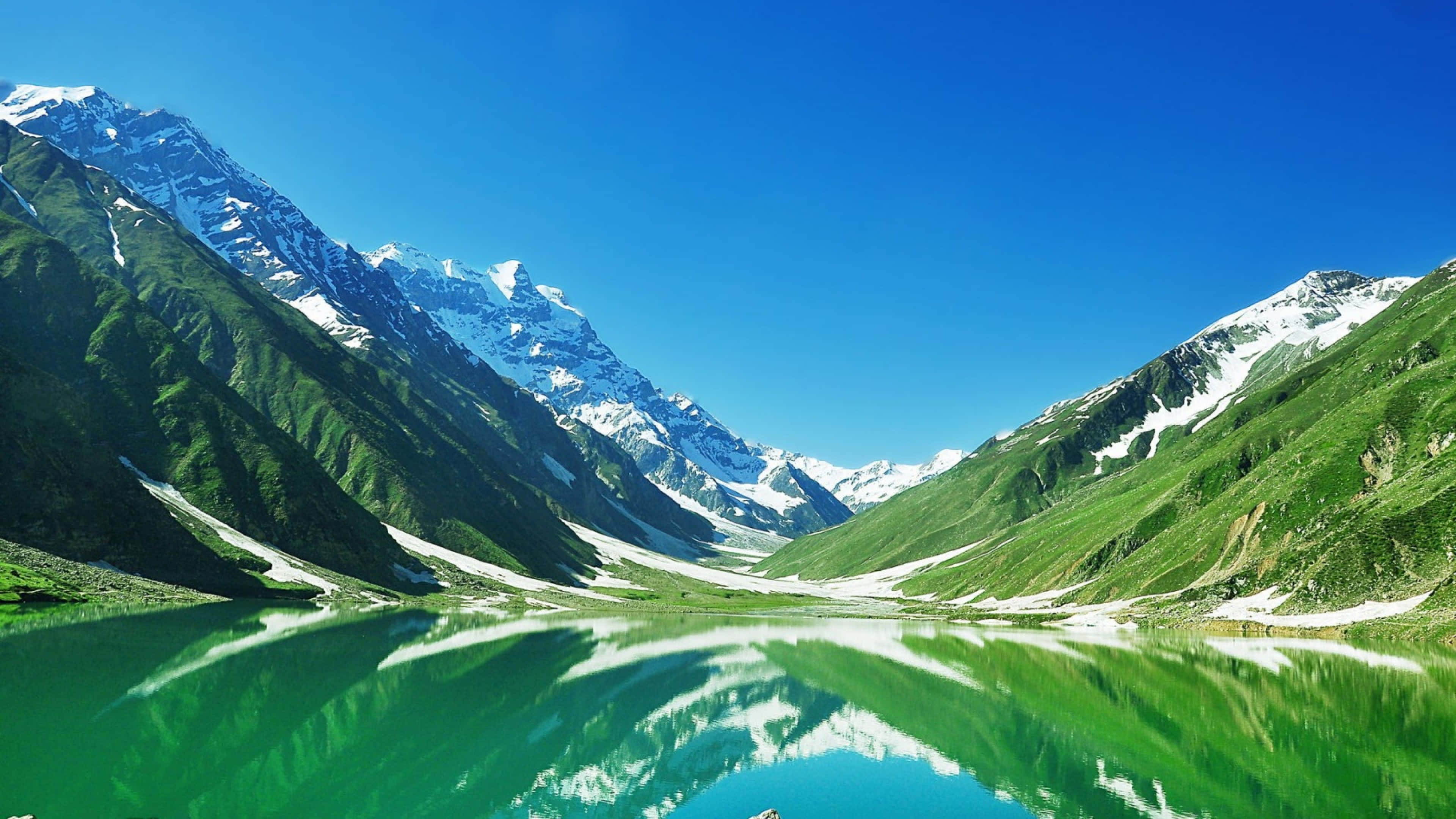 Pakistan 桌布 壁紙 — 寧靜的高山湖泊映照著白雪覆蓋的山峰，藍天白雲下綠草如茵的山坡。