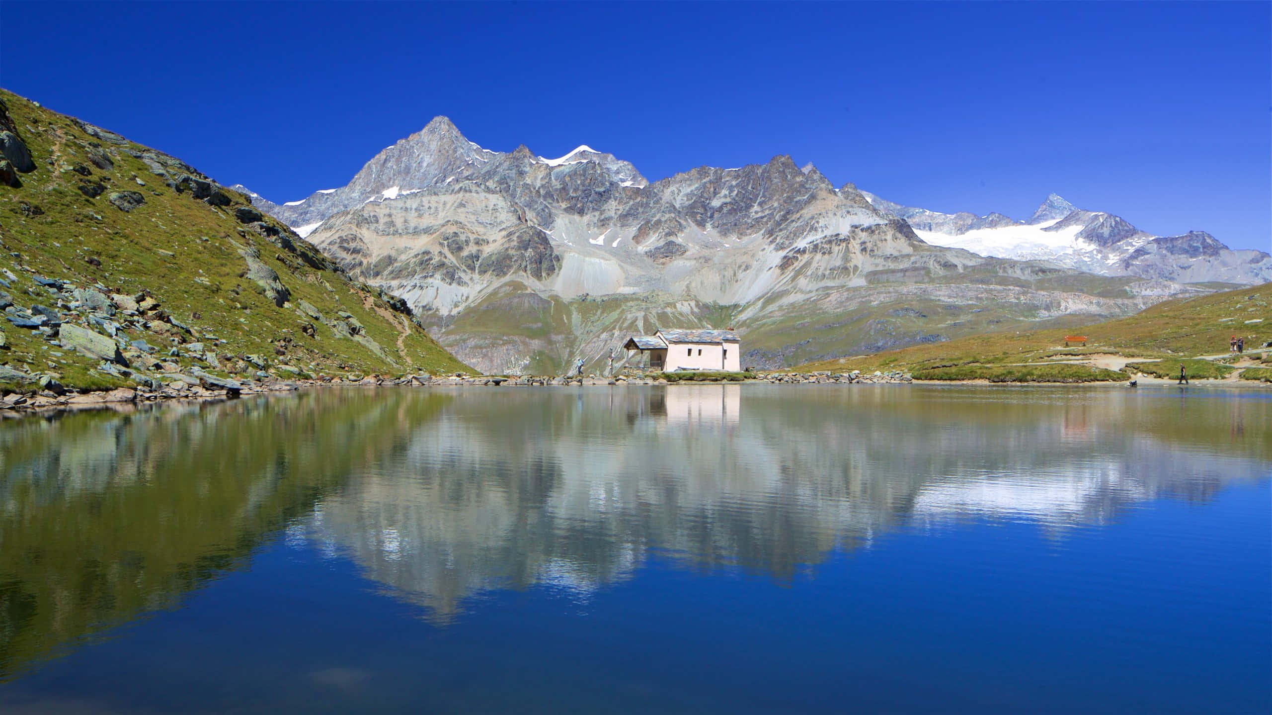 Switzerland 桌布 壁紙 — 寧靜的高山湖泊映照著白雪覆蓋的山峰和一座孤獨的石造教堂，在清澈的藍天下。