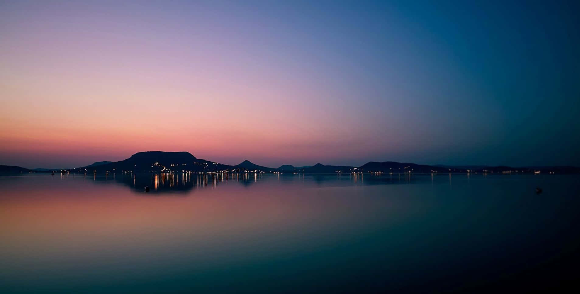 Evening 桌布 壁紙 — 寧靜的黃昏場景，平靜的湖面倒映著粉紅和紫色的天空，遠處的山巒輪廓點綴著城市燈光。