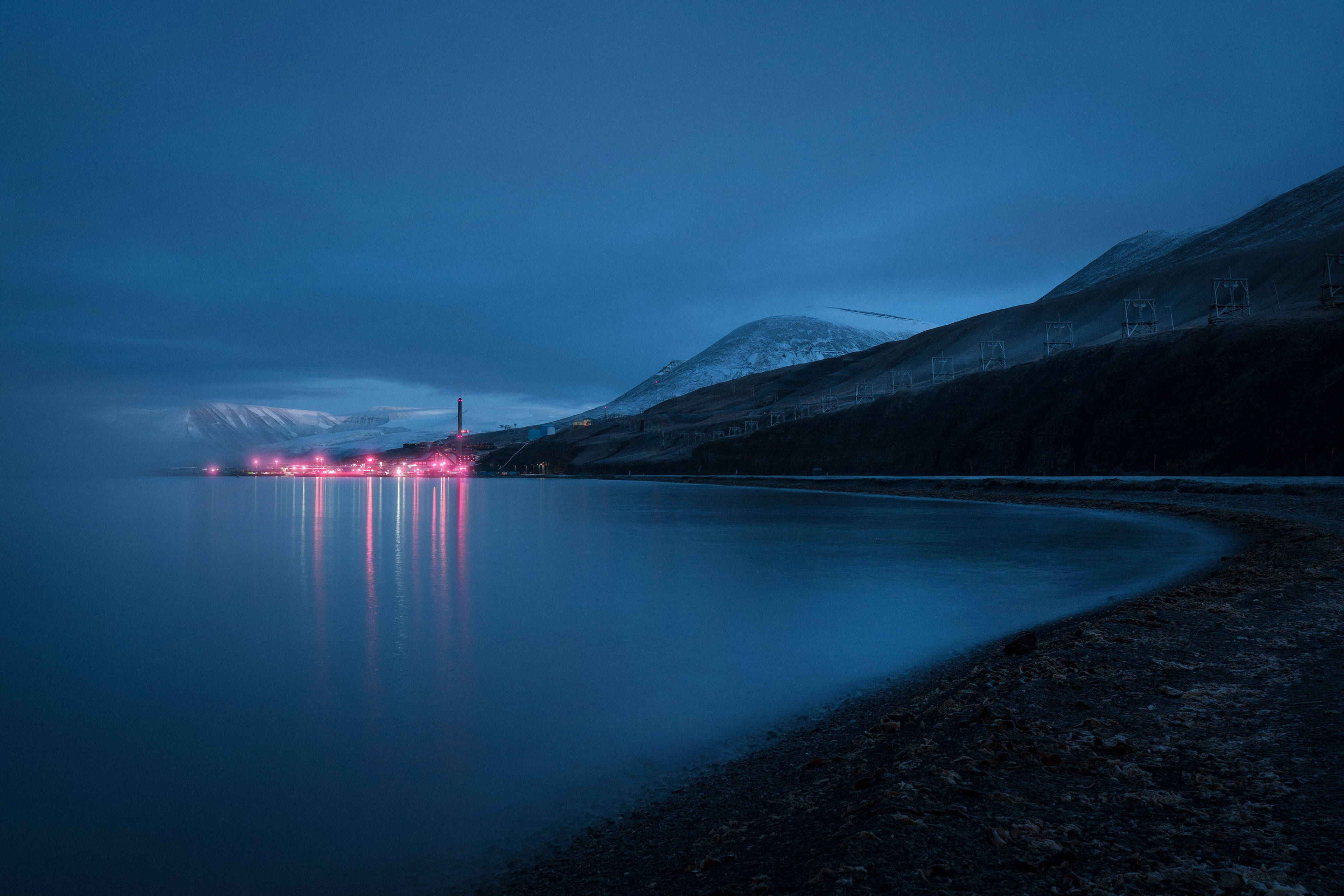 Svalbard 桌布 壁紙 — 寧靜的黃昏海岸景色，發光的紅燈映照在平靜的水面上，旁邊是白雪覆蓋的山脈。