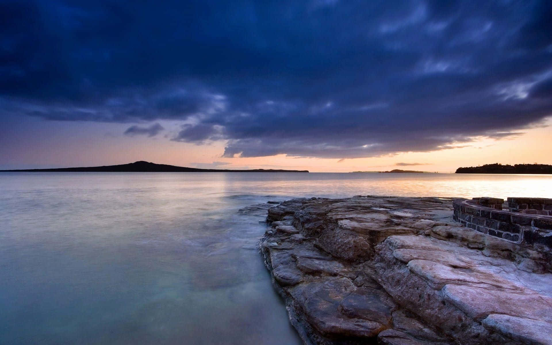 Soothing 桌布 壁紙 — 寧靜的黃昏海景，平滑的水流經過崎嶇的石崖，天空呈現戲劇性的紫色和橙色。
