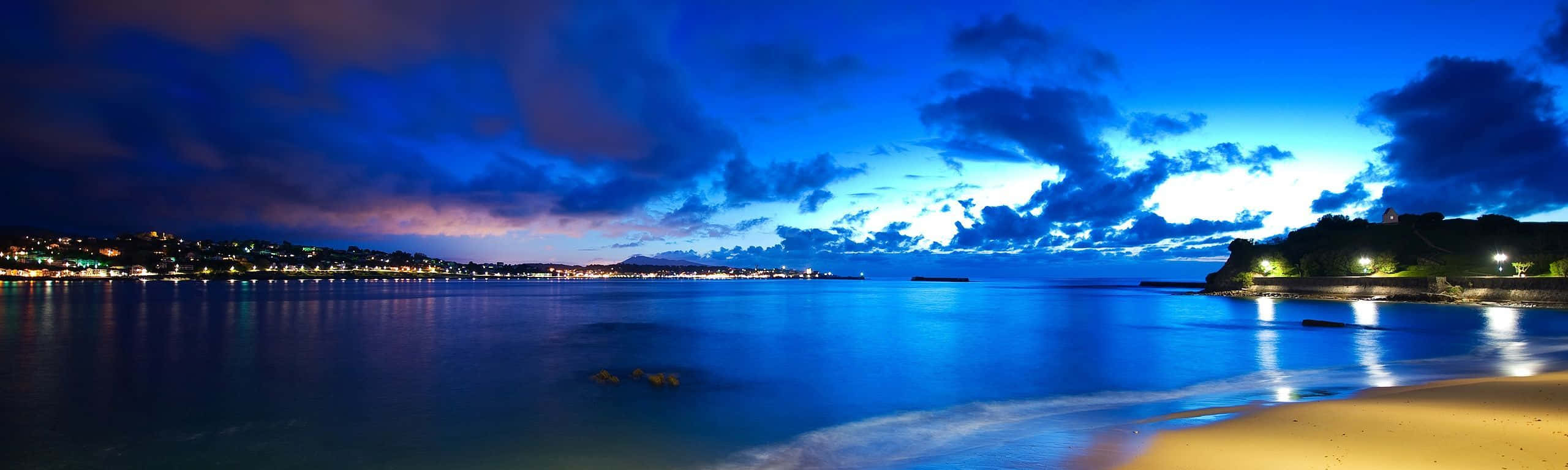 wide 桌布 壁紙 — 寧靜的黃昏海灘全景，在戲劇性的藍天下，閃爍的城市燈光倒映在平靜的水面上。