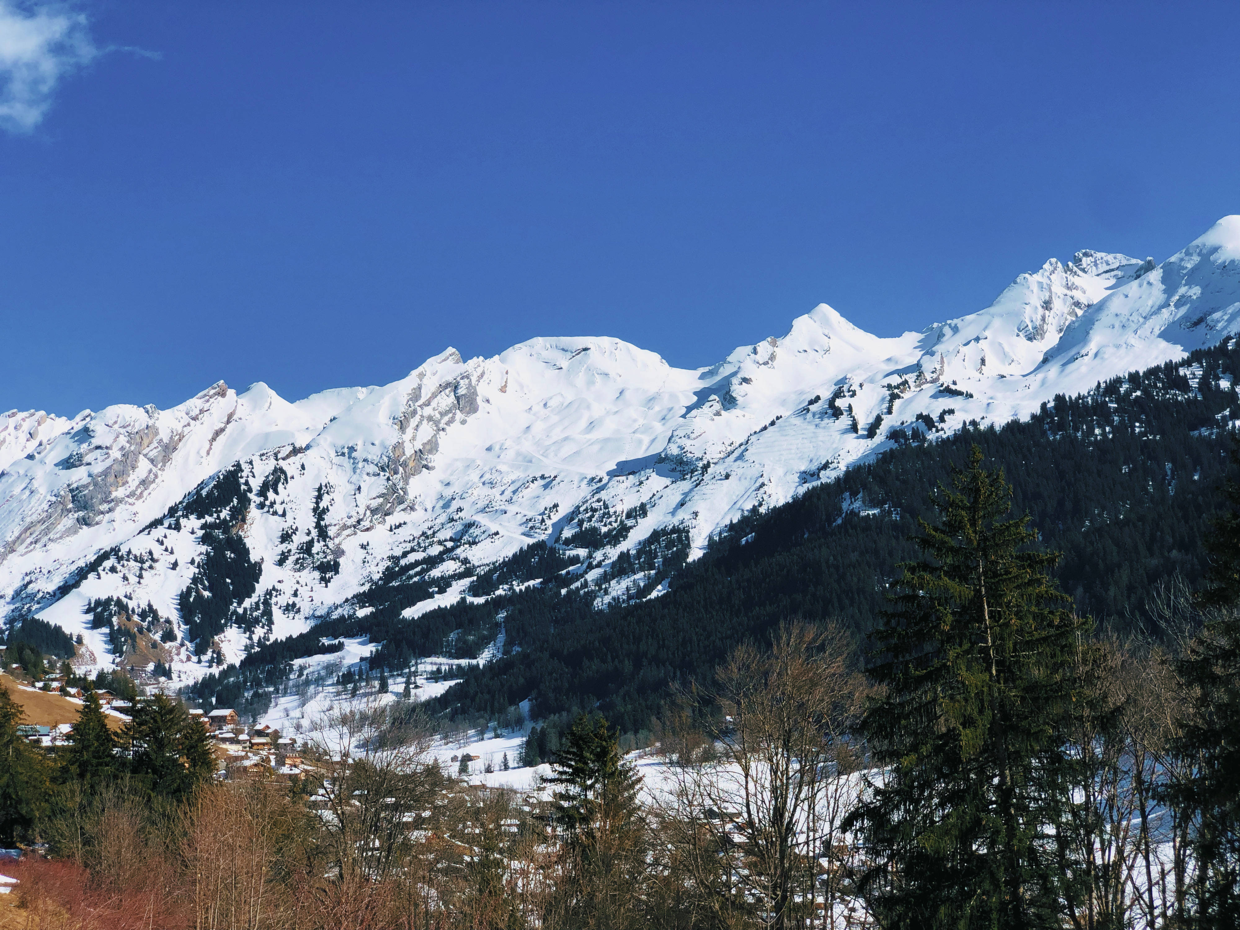 寧靜雪峰山脈 壁紙 — 白雪覆蓋的山峰在晴朗的藍天下，聳立於森林覆蓋的山谷之上，山谷中散落著房屋。