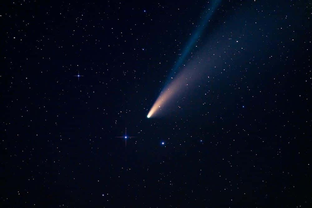 寬85英里的彗星影像 壁紙 — 一顆明亮的彗星劃過滿天星斗的夜空，留下一條發光的藍色和橙色尾巴。