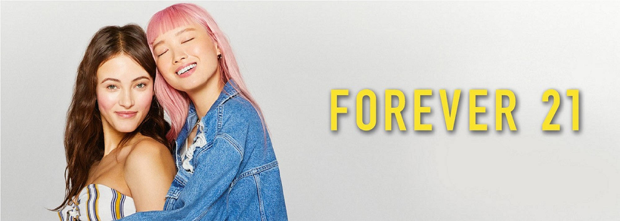 寬螢幕Forever 21時尚海報 壁紙 — 兩位微笑的年輕女性，一位粉紅色頭髮靠著另一位，背景是純色帶有黃色Forever 21文字。