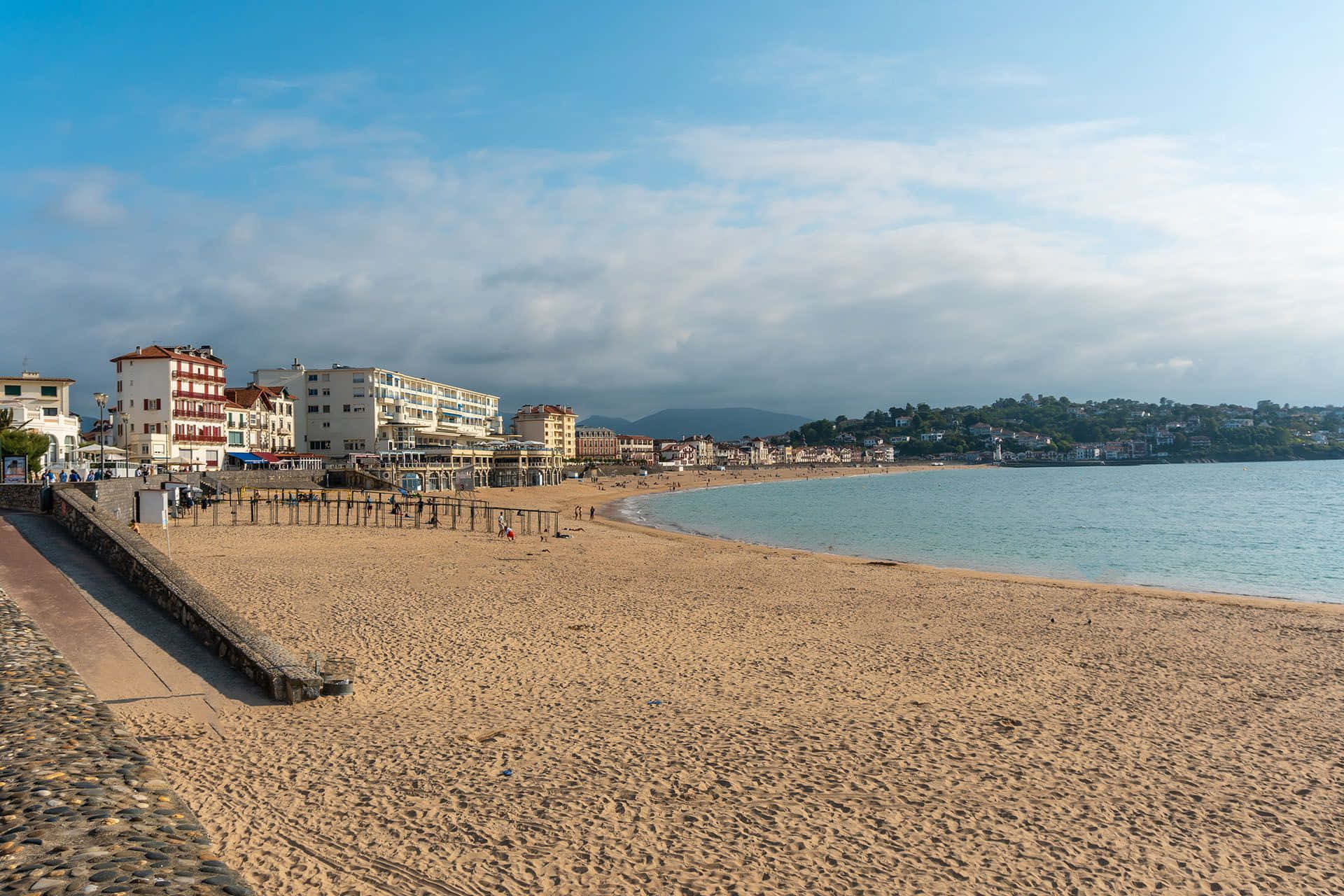 French Beach 桌布 壁紙 — 寬闊的沙灘沿著平靜的碧綠海水蜿蜒，色彩繽紛的海濱建築在部分多雲的藍天下。
