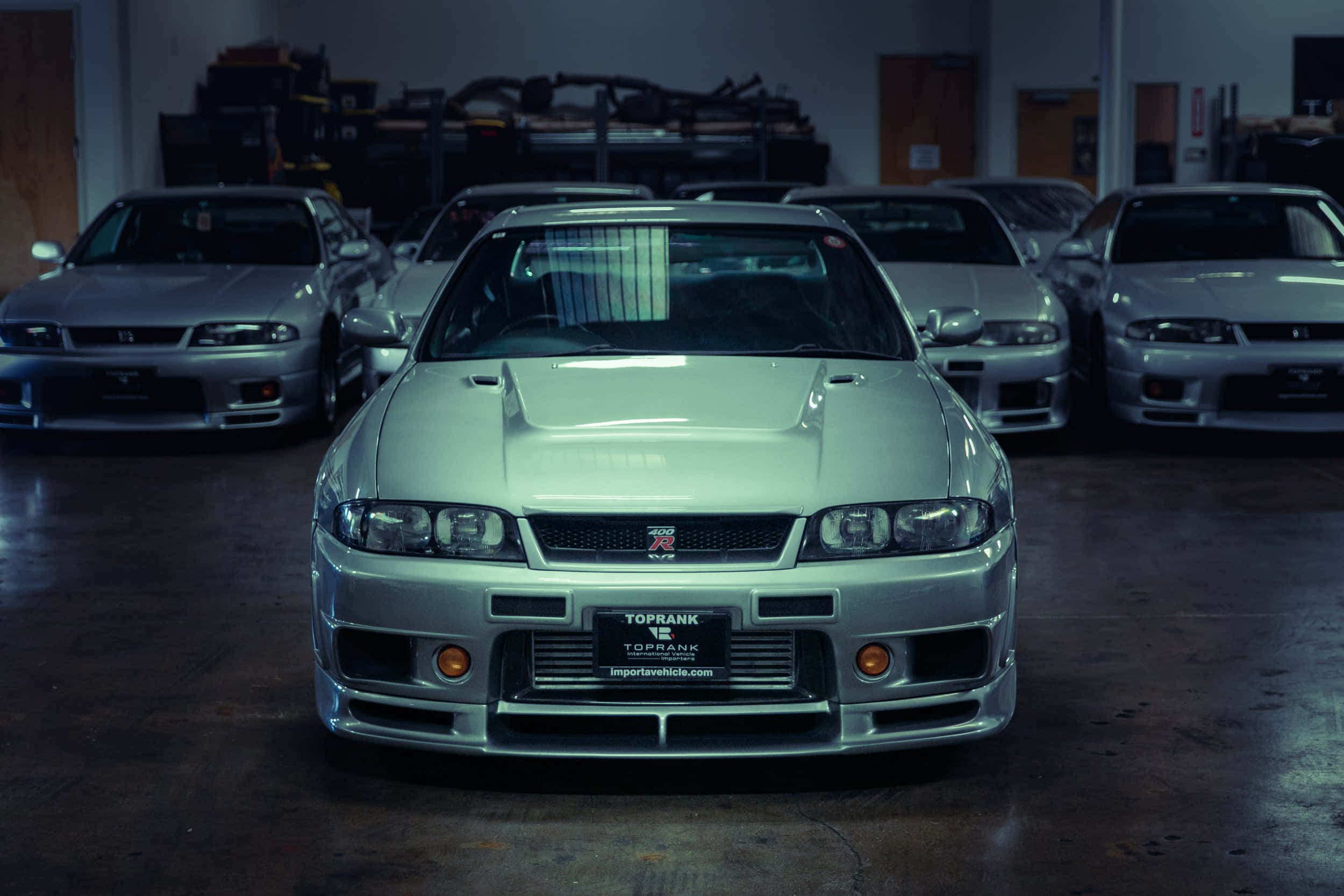 寬體版 R33 GTR，強勁如神獸 壁紙 — 一架銀色日產Skyline R34 GT-R喺昏暗嘅車庫入面，被其他經典車包圍，面向前方。