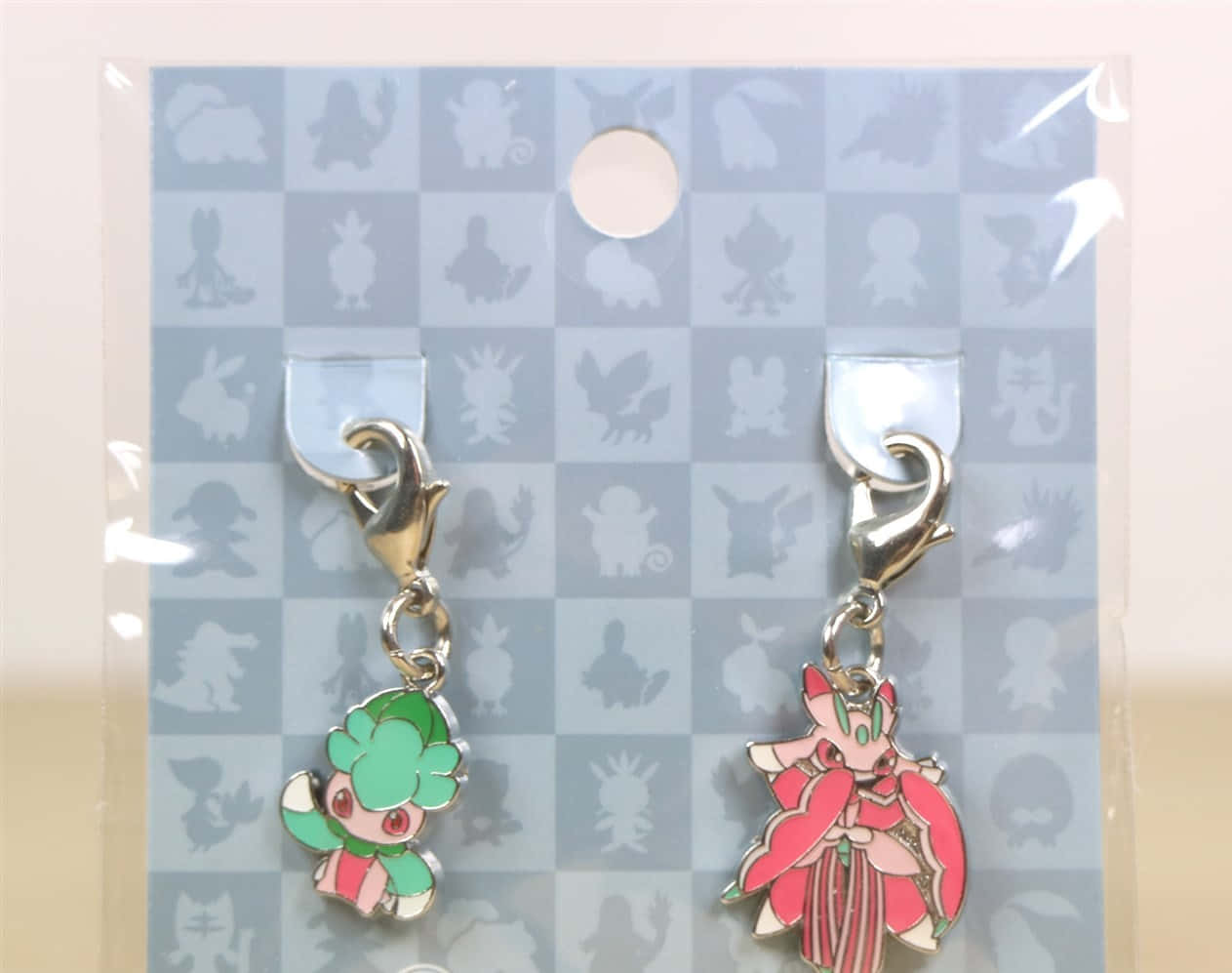 寶可夢 Fomantis 與 Lurantis 鑰匙圈 壁紙 — 兩個色彩繽紛的寵物小精靈琺瑯吊飾用銀色夾子掛在帶有模糊寵物小精靈剪影的格子展示卡上。