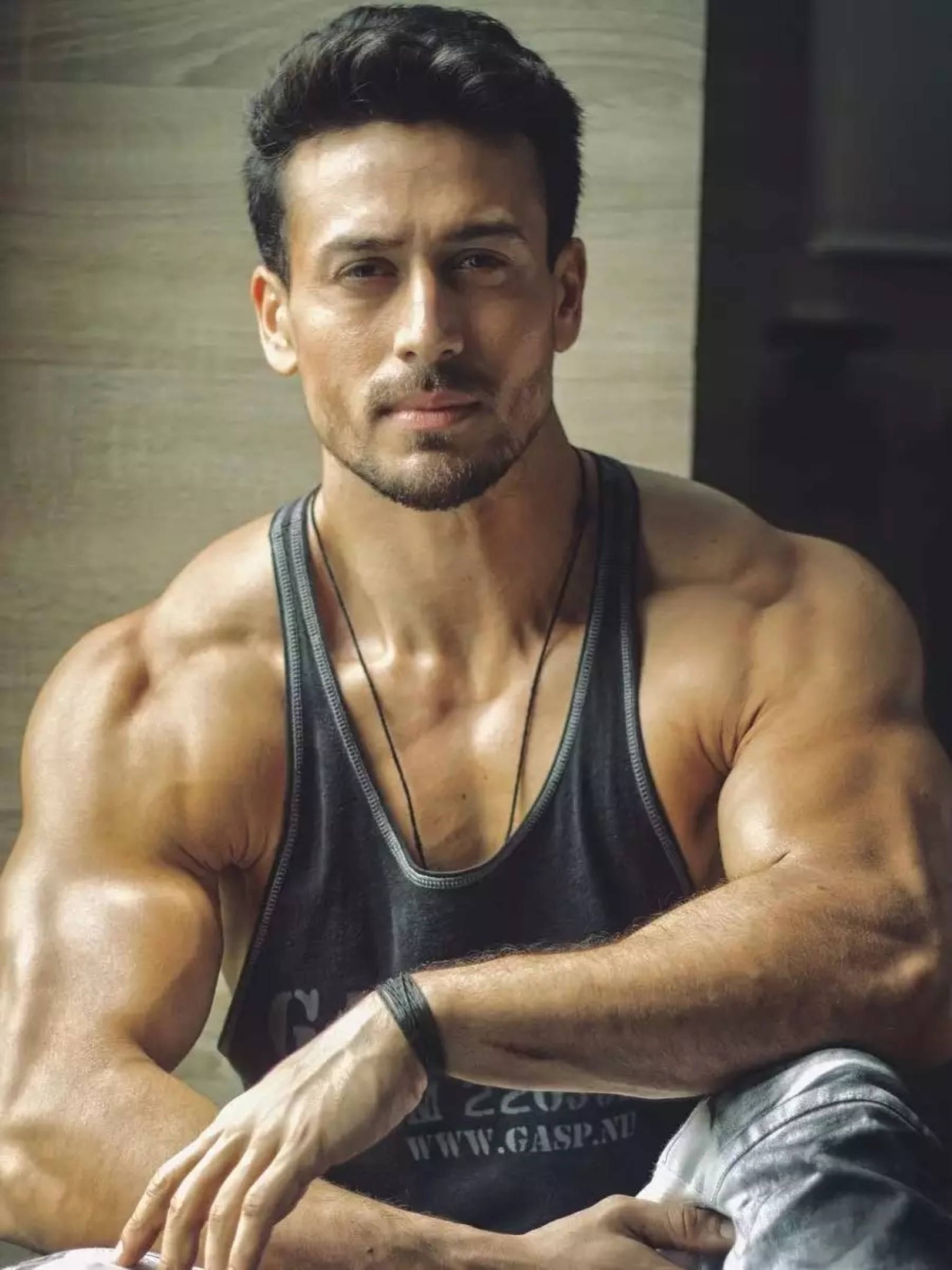 寶萊塢 Tiger Shroff 壁紙 — 一個肌肉發達、黑髮帶鬍渣的男人自信地凝視鏡頭，身穿黑色背心。
