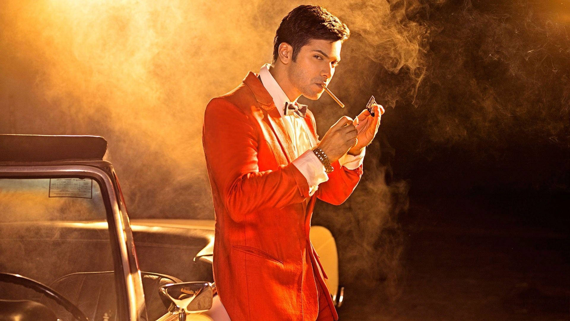 寶萊塢明星 Varun Dhawan 壁紙 — 一名身穿紅色西裝的男子在金色煙霧和戲劇性燈光下，於一輛復古車旁吸煙。