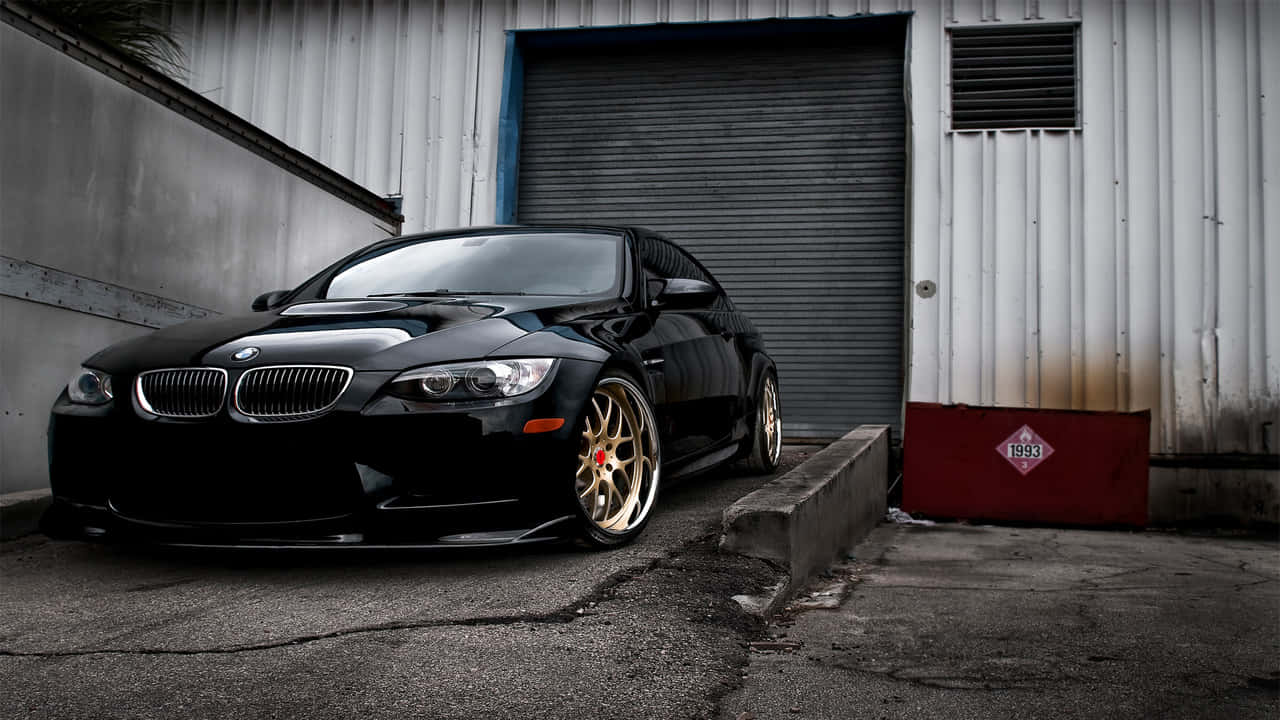 寶馬豪華車停於城市天際線前 壁紙 — 一輛閃亮黑色BMW M3雙門跑車配金色輪圈，停在風化的工業車庫門前。