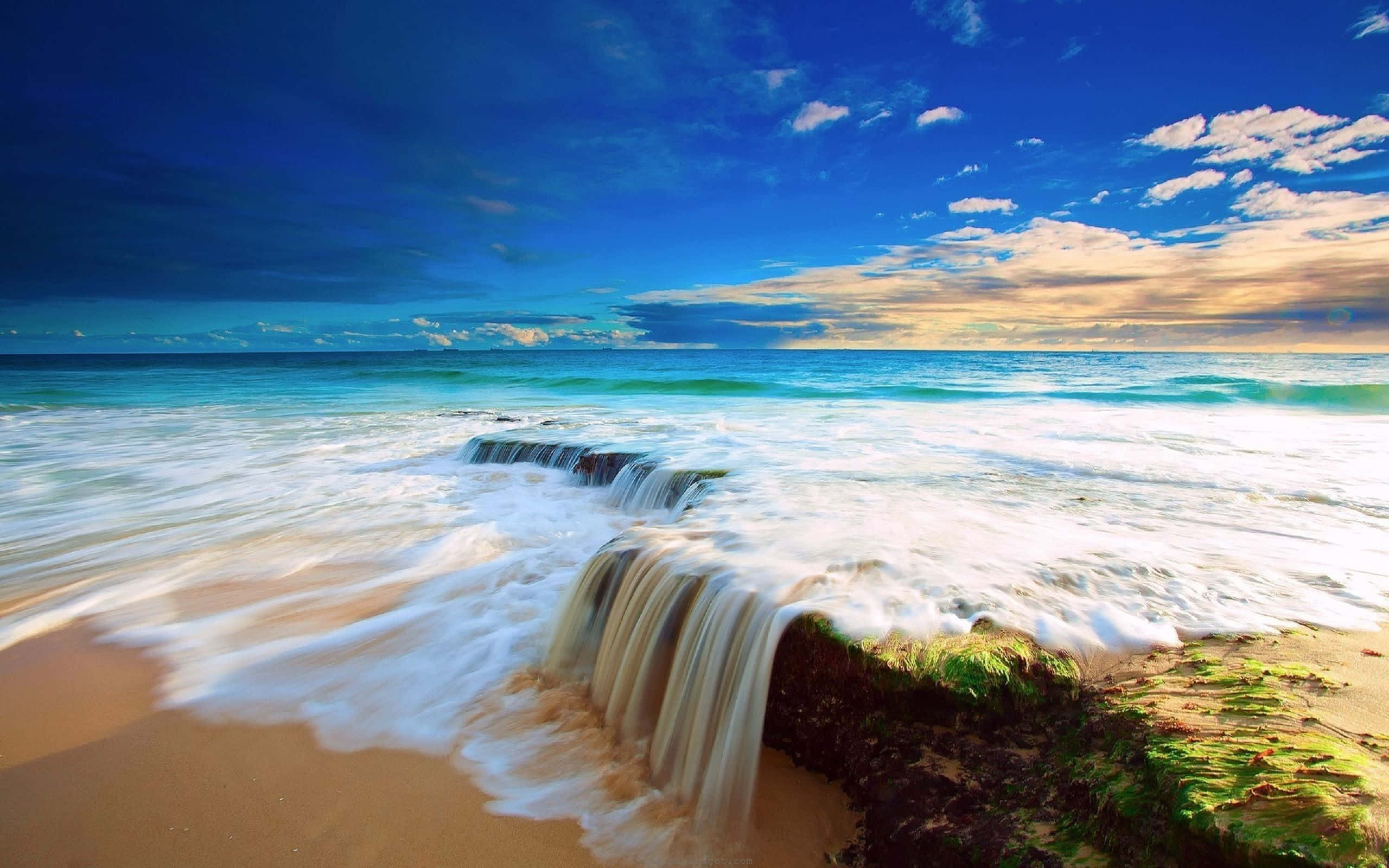 將夢想變為現實的寧靜海灘主題背景 壁紙 — 日落時分，海浪如瀑布般越過岩石露頭，灑落在金色沙灘上，天空湛藍，雲朵點綴。