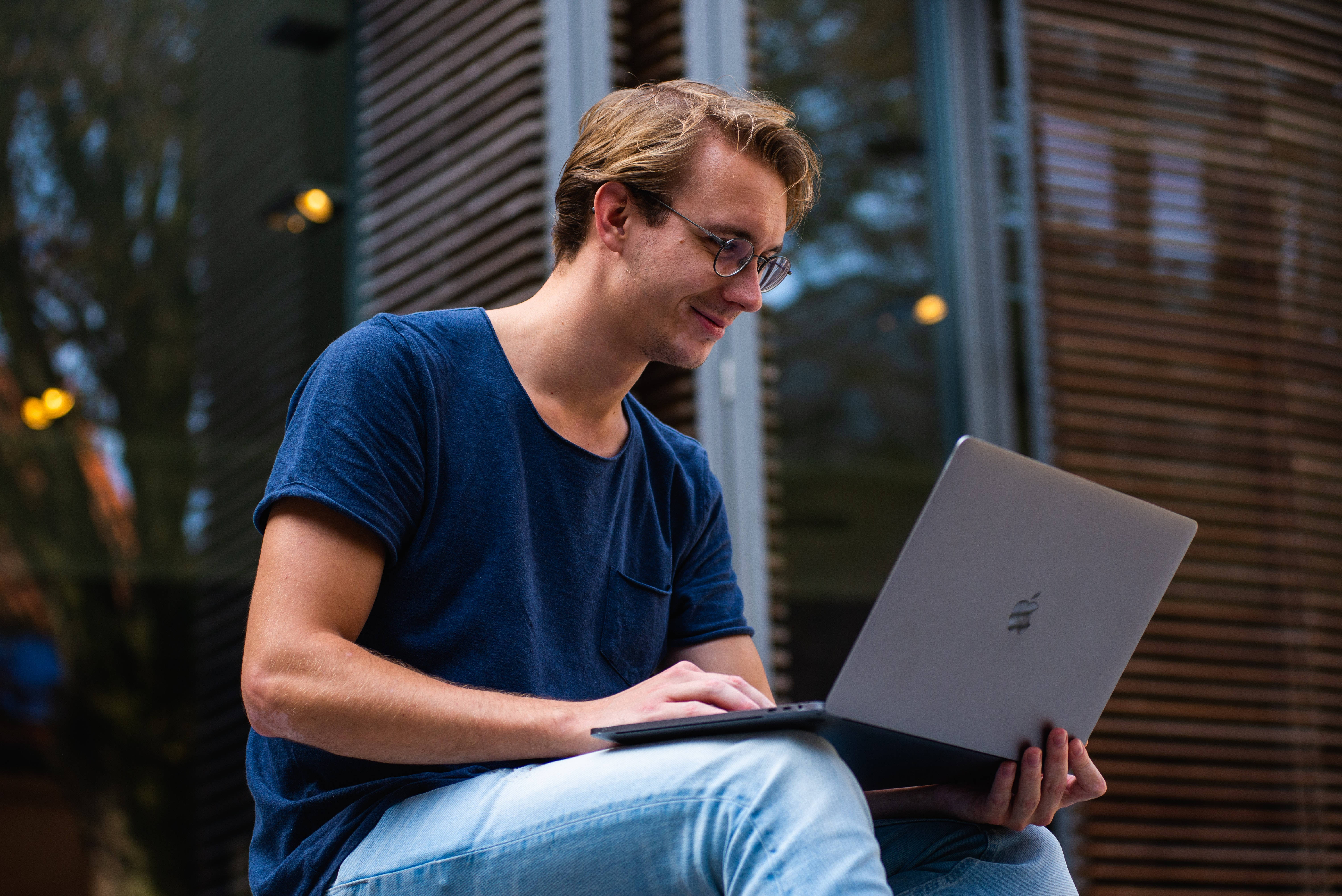 專業人士使用MacBook工作 壁紙 — 一位戴眼鏡的年輕男子在戶外靠近木條牆的地方微笑著在銀色筆記本電腦上打字。