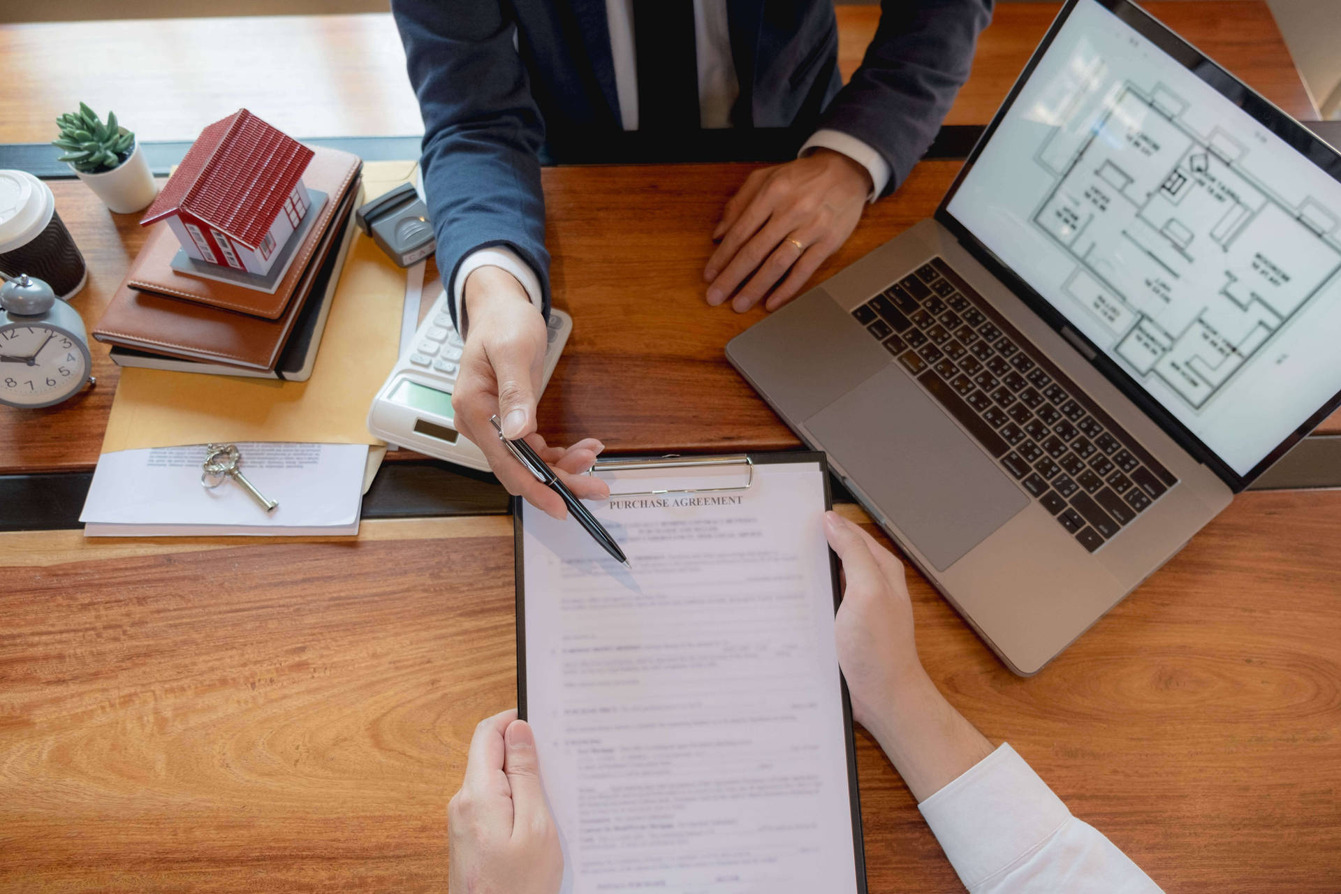 Two people signing a real estate purchase agreement at a wooden desk with laptop and keys.