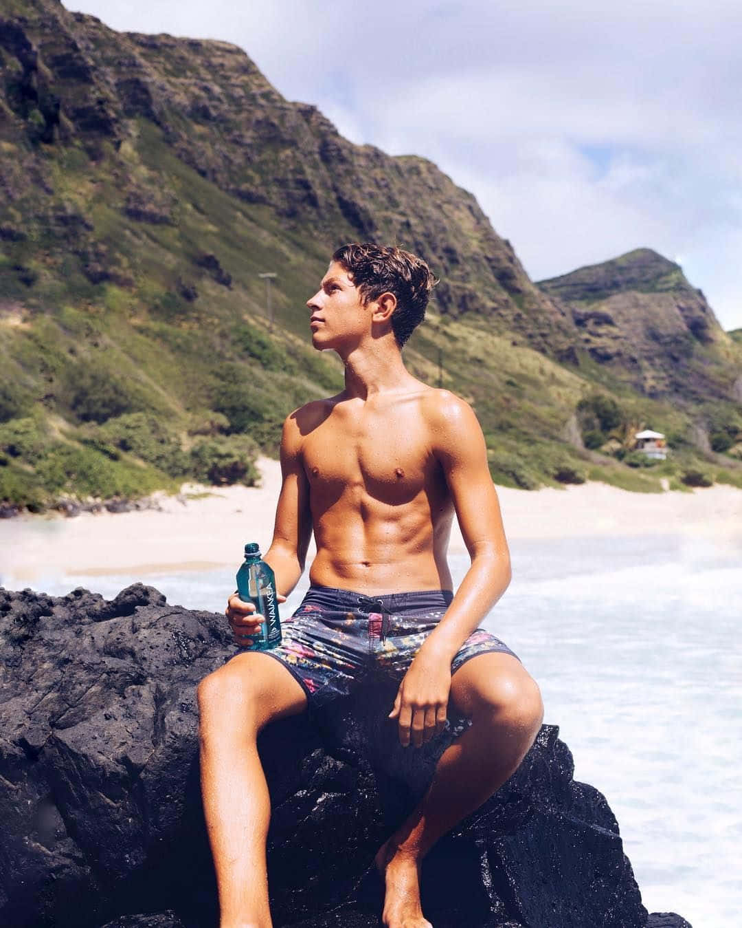 一個肌肉發達的男人坐在海邊的黑色岩石上，手持水樽。