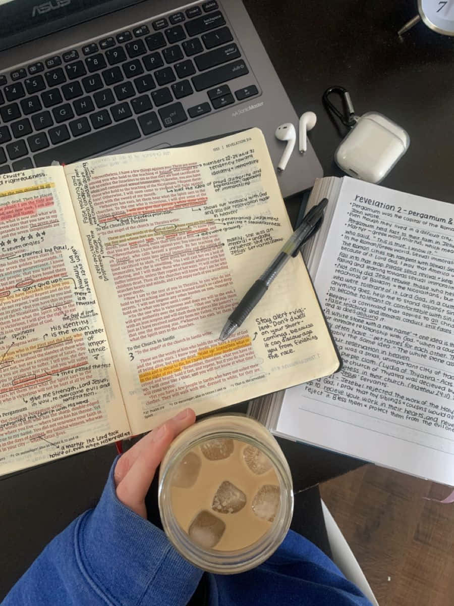 專注學習：學生在書房中 壁紙 — 一隻手拿著冰咖啡，旁邊放著打開的聖經和手提電腦，在木桌上，還有耳塞和筆。