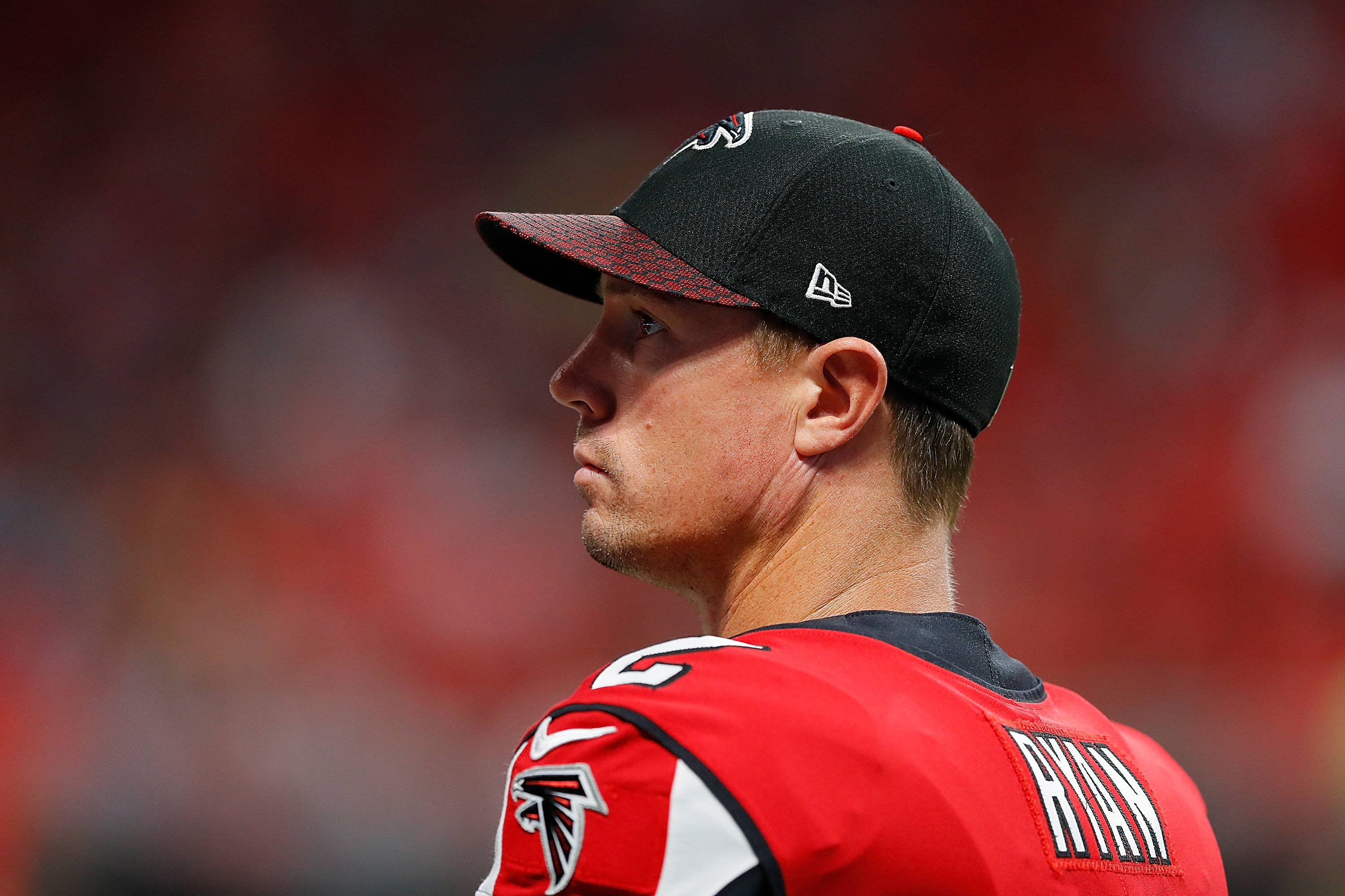 Matt Ryan 桌布 壁紙 — 專注的亞特蘭大獵鷹美式足球員身穿紅色球衣和黑色帽子，背景是模糊的體育場人群的 close-up 側面照。
