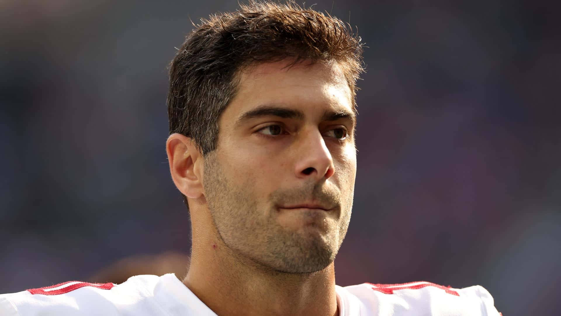 Jimmy Garoppolo 桌布 壁紙 — 專注的足球運動員身穿白色球衣配紅色飾邊，在明亮的日光下望向鏡頭外的特寫肖像。