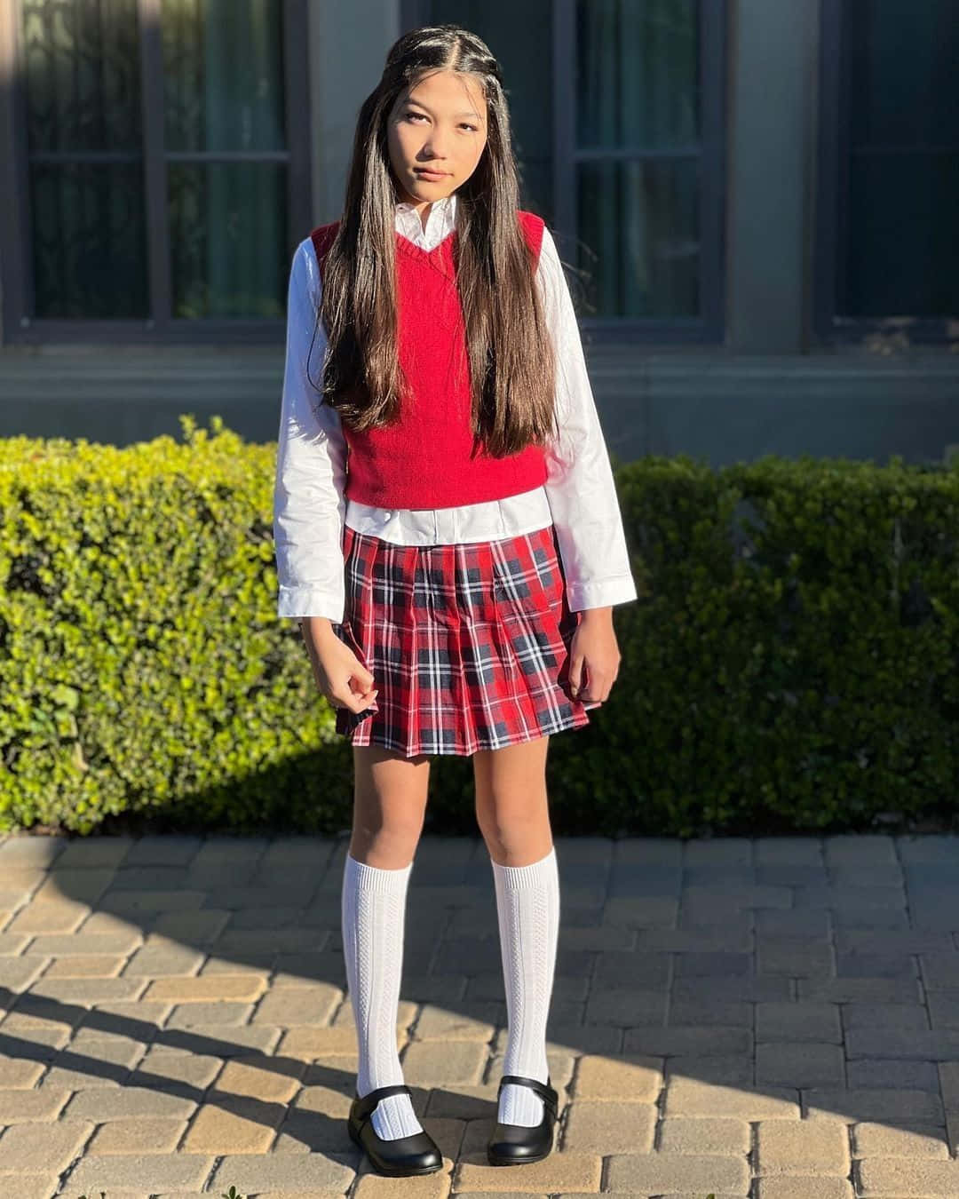 穿著校服的女孩站在磚砌小徑上，旁邊有綠色樹籬