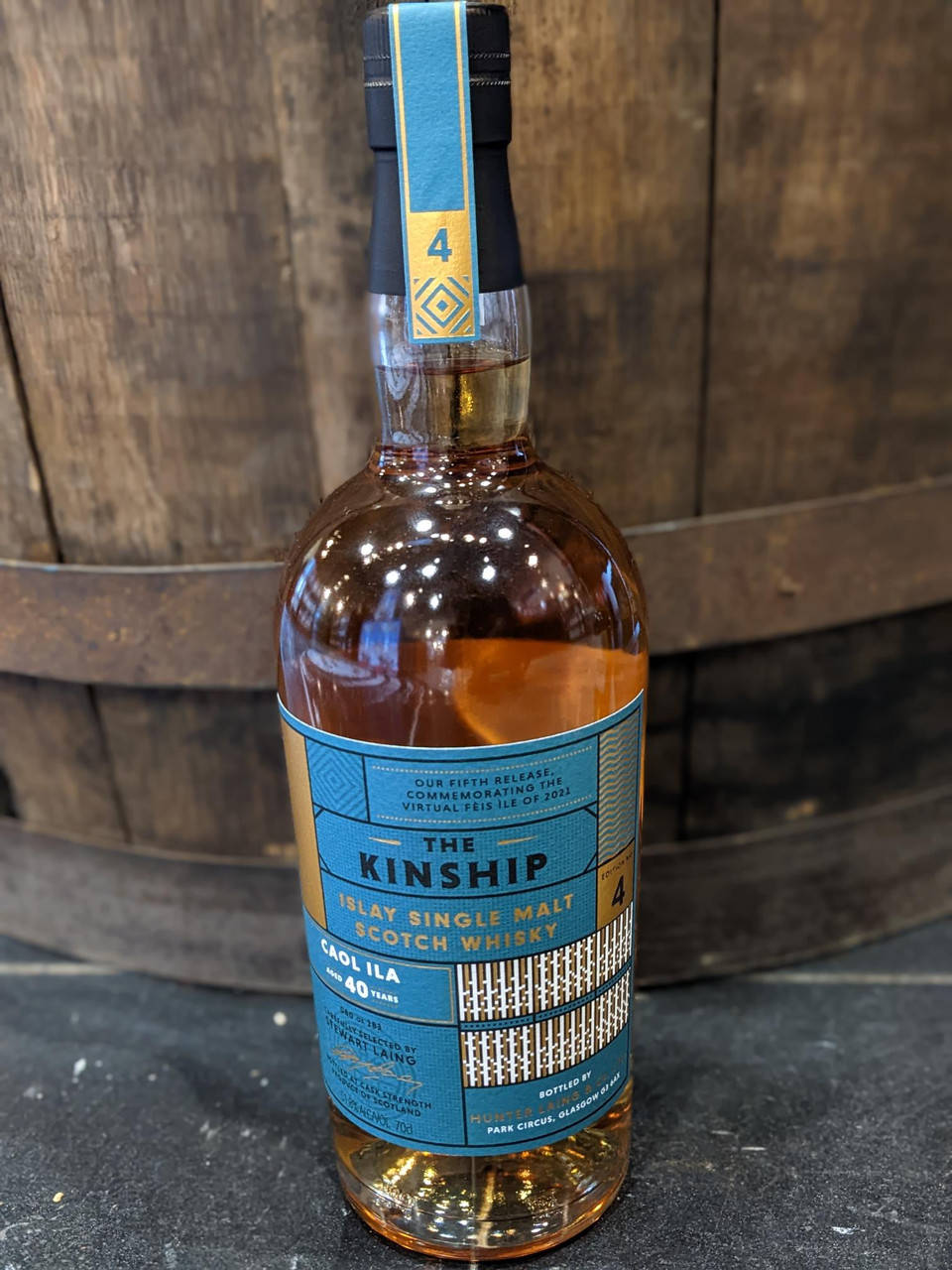 一瓶帶有藍色標籤的 Kinship 威士忌靠在木桶前。